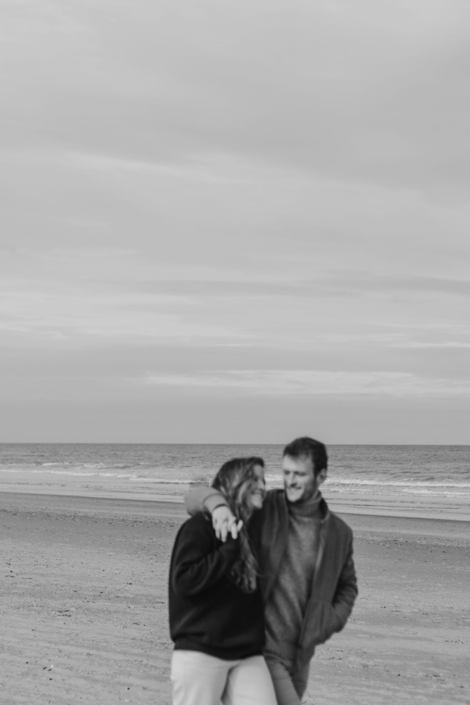A couple embraces while walking along a misty beach shore with cloudy skies in black and white.