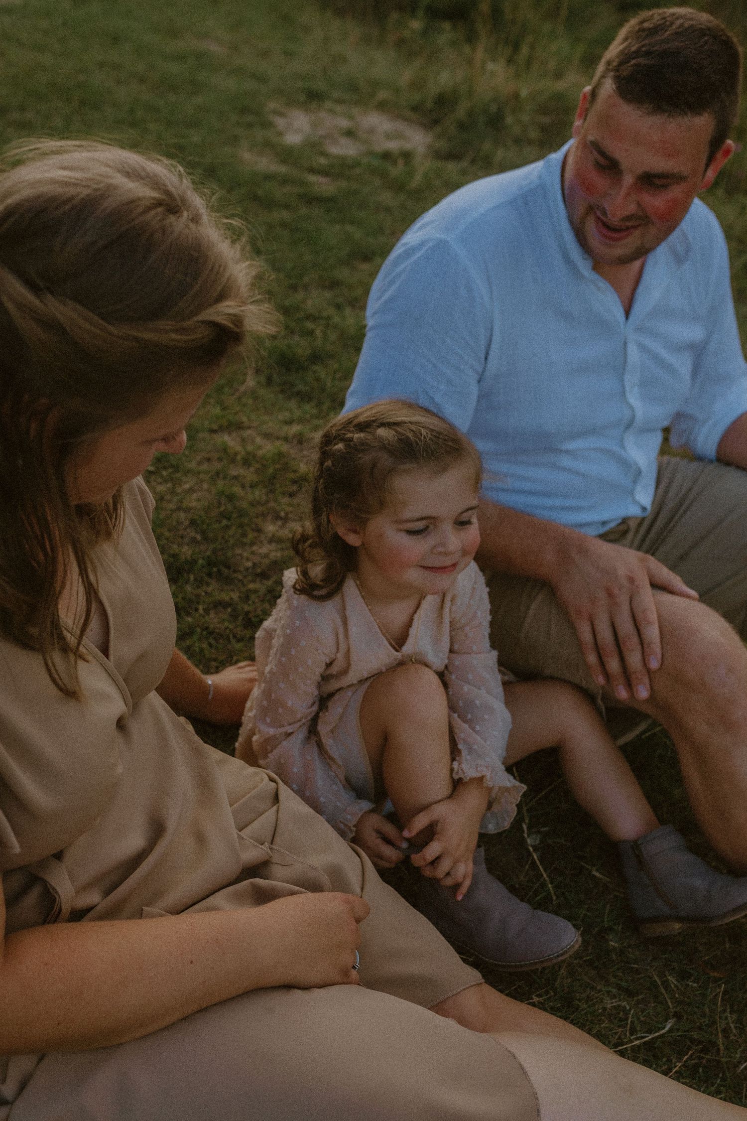 A family sits together outdoors on grass during golden hour, sharing a tender moment in casual attire.