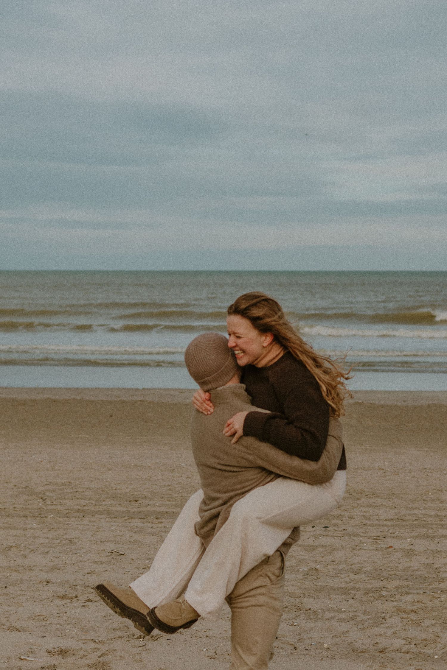 A couple embraces playfully on a cloudy beach with gray waves and overcast skies in the background.