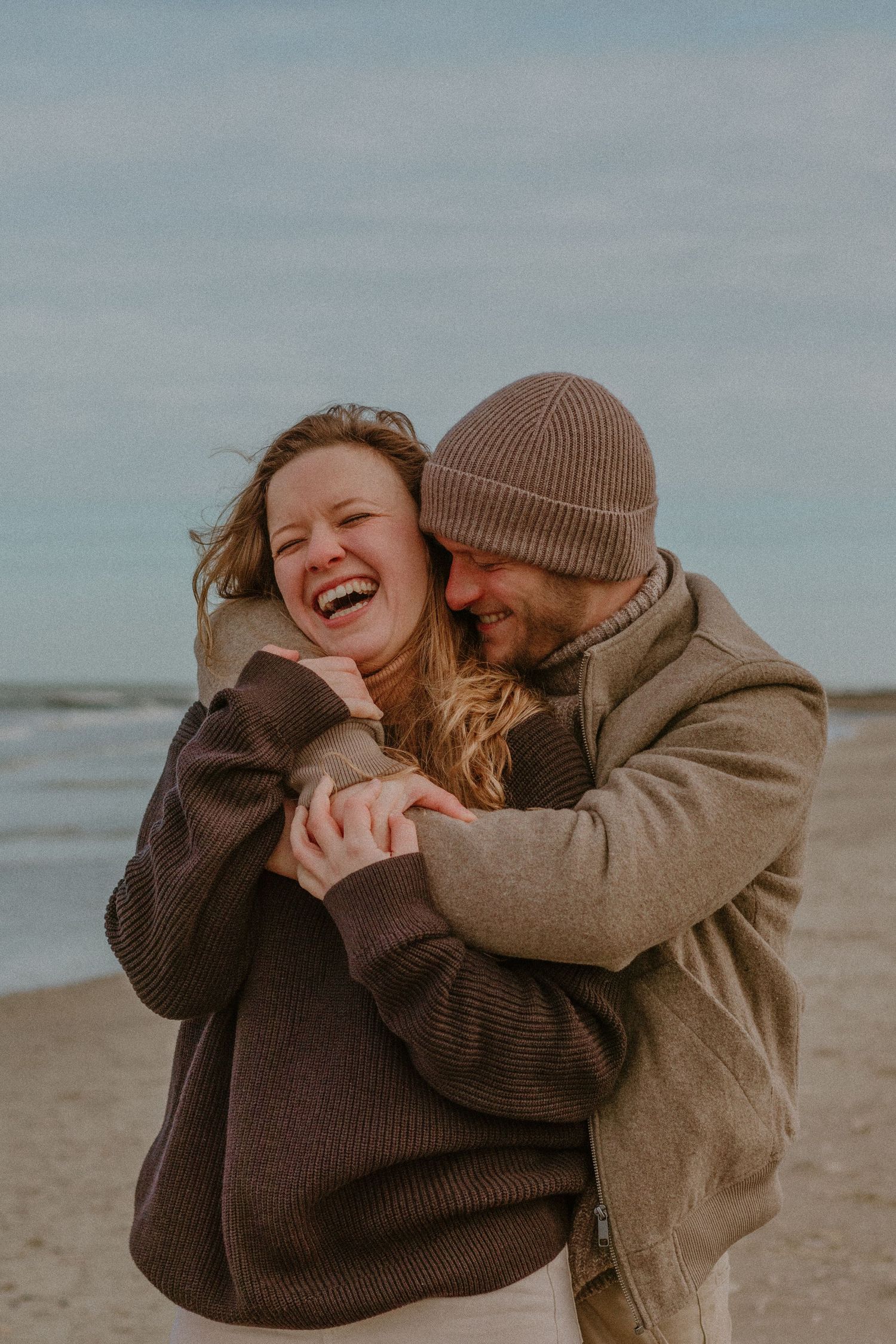 A joyful couple in cozy sweaters and beanies embrace and laugh together on a cloudy beach.