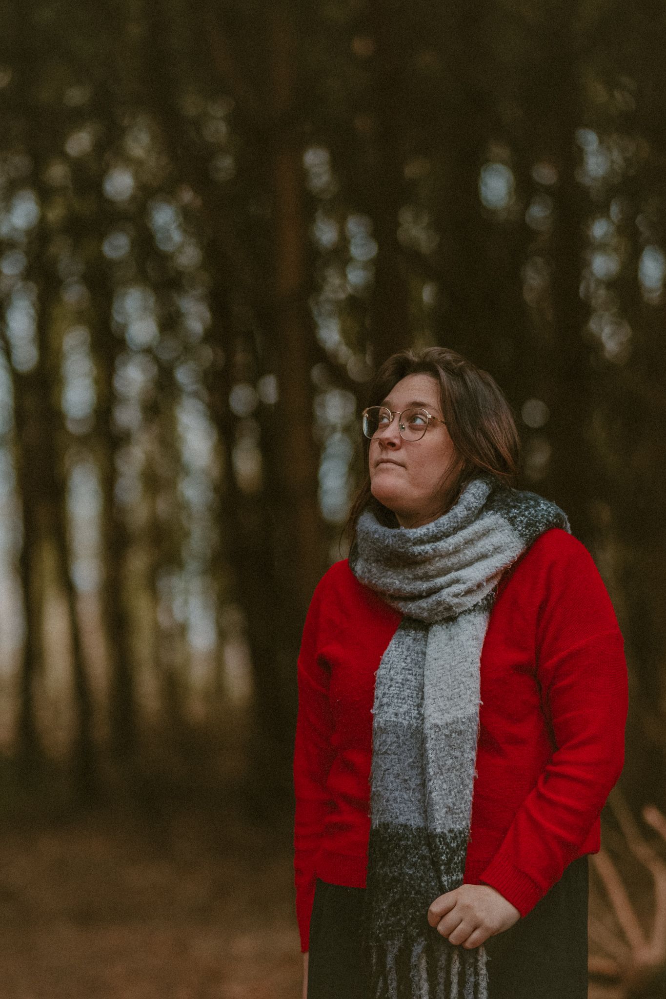 A person in a bright red sweater and gray scarf poses thoughtfully in a moody forest setting during autumn.
