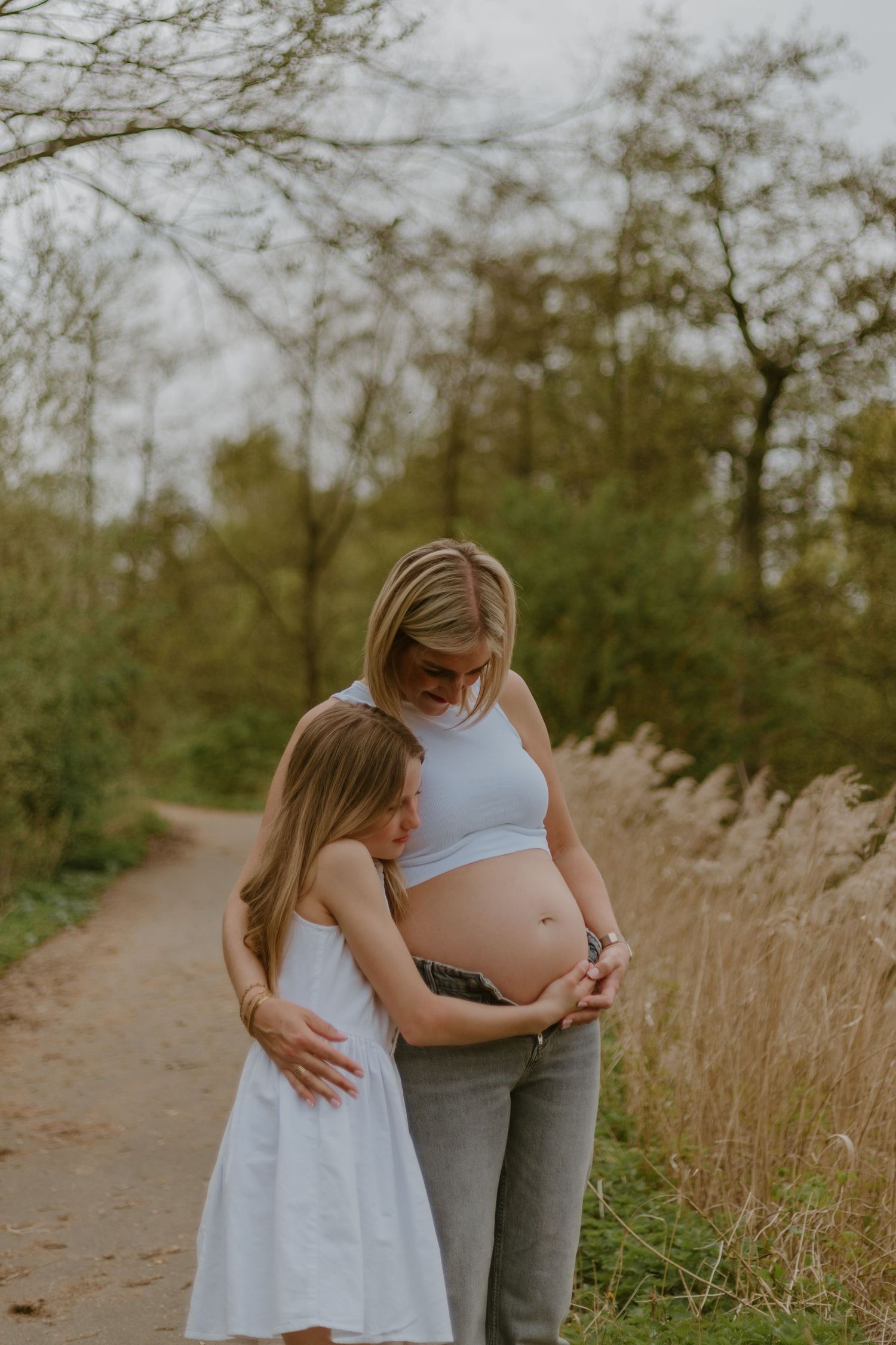 A young girl in a white dress hugs her pregnant mother's baby bump on a scenic outdoor path surrounded by trees.