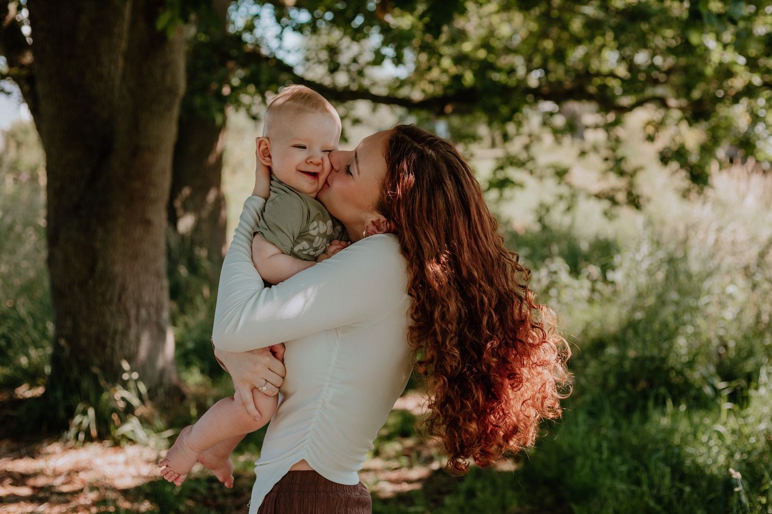 A mother with long red hair kisses her smiling baby outdoors near a large tree in natural sunlight.