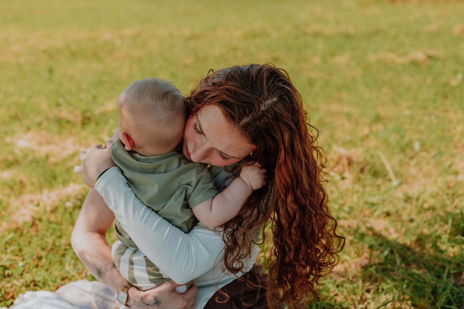A mother with curly red hair tenderly hugs her baby outdoors on green grass in warm sunlight.