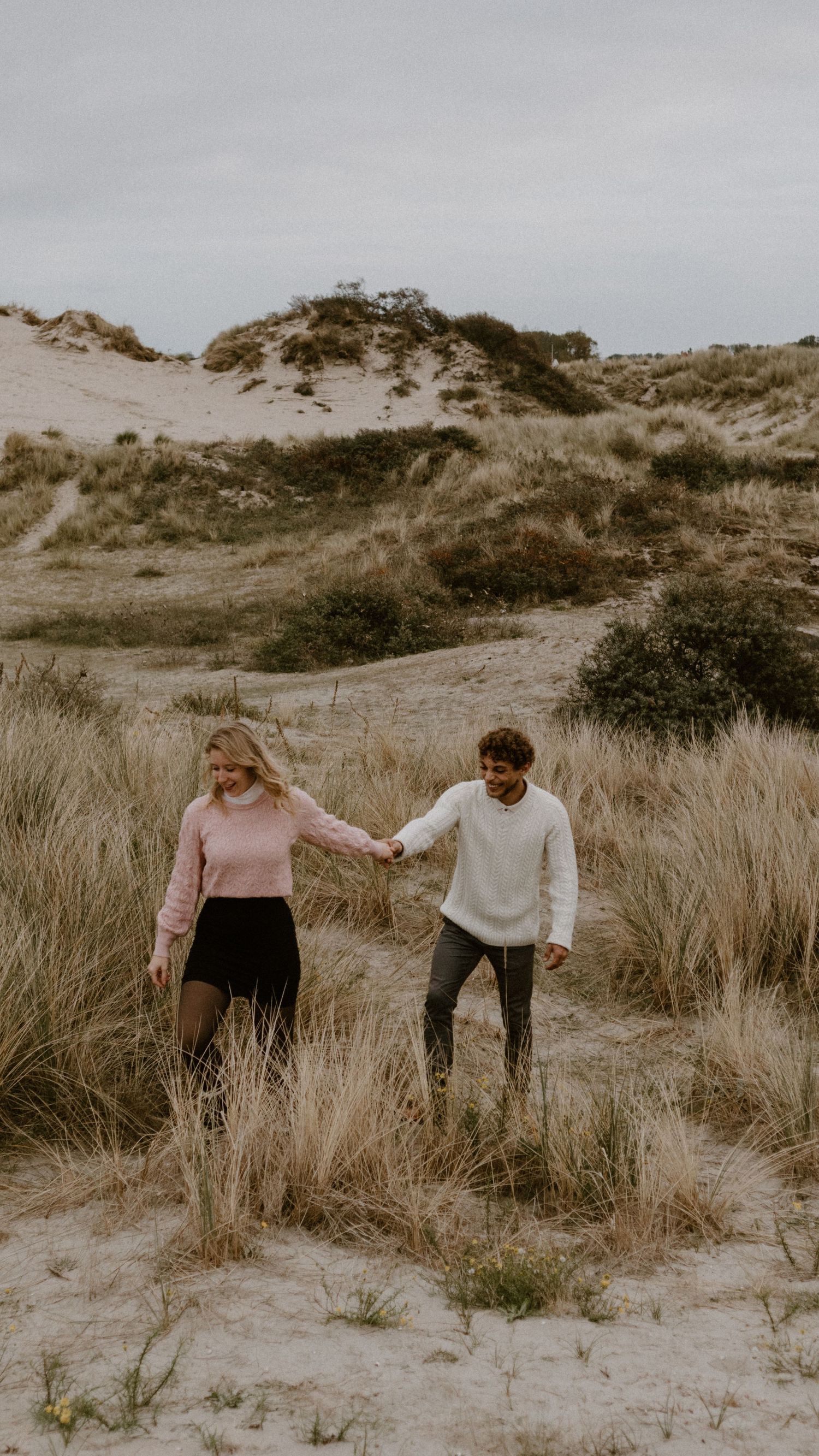Een stel houdt elkaars hand vast terwijl ze door de kustduinen met hoog gras wandelen.