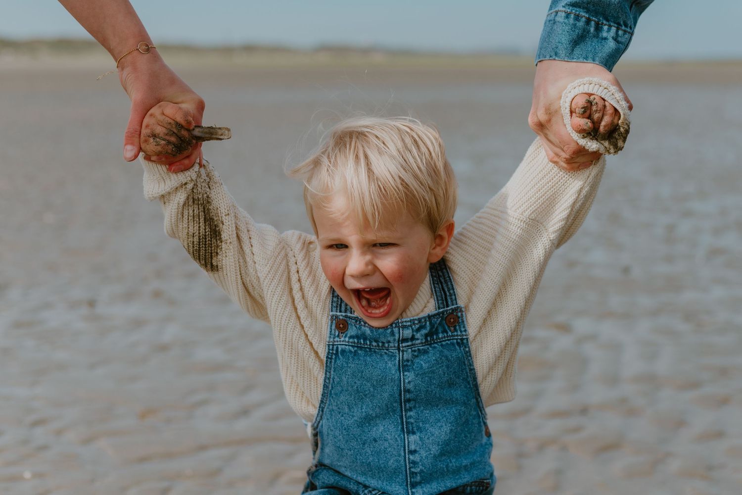 Joyful toddler in denim overalls laughing on a sandy beach, held up by adults hands on a cloudy day.
