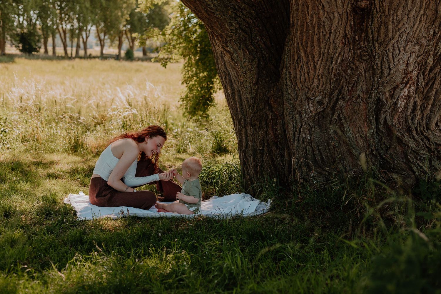 Mother and baby sitting on white blanket under large tree in sunny green park during outdoor family picnic.