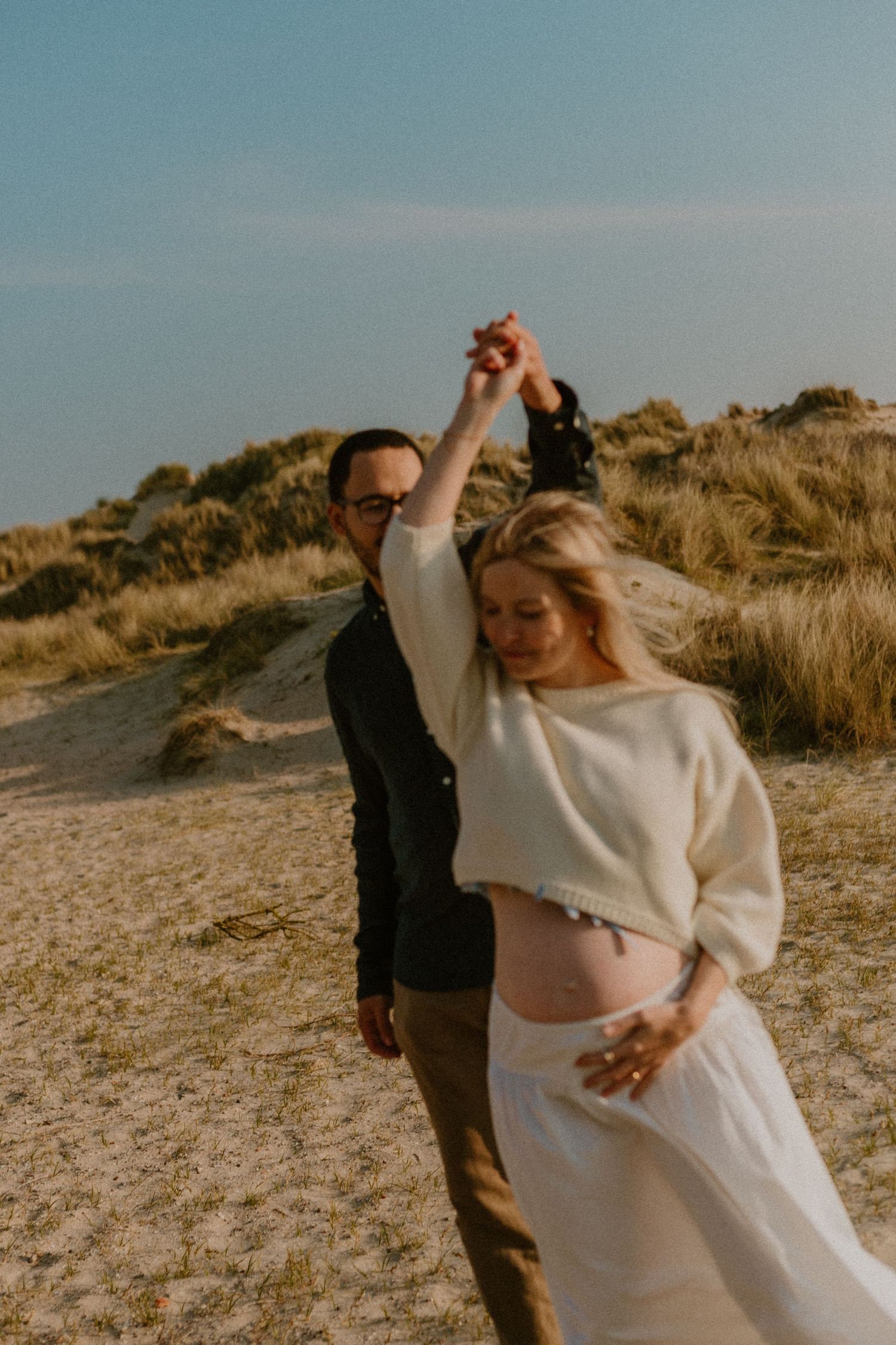Pregnant couple dancing on sandy beach dunes, woman in white crop top holding baby bump, man in dark jacket, golden hour.