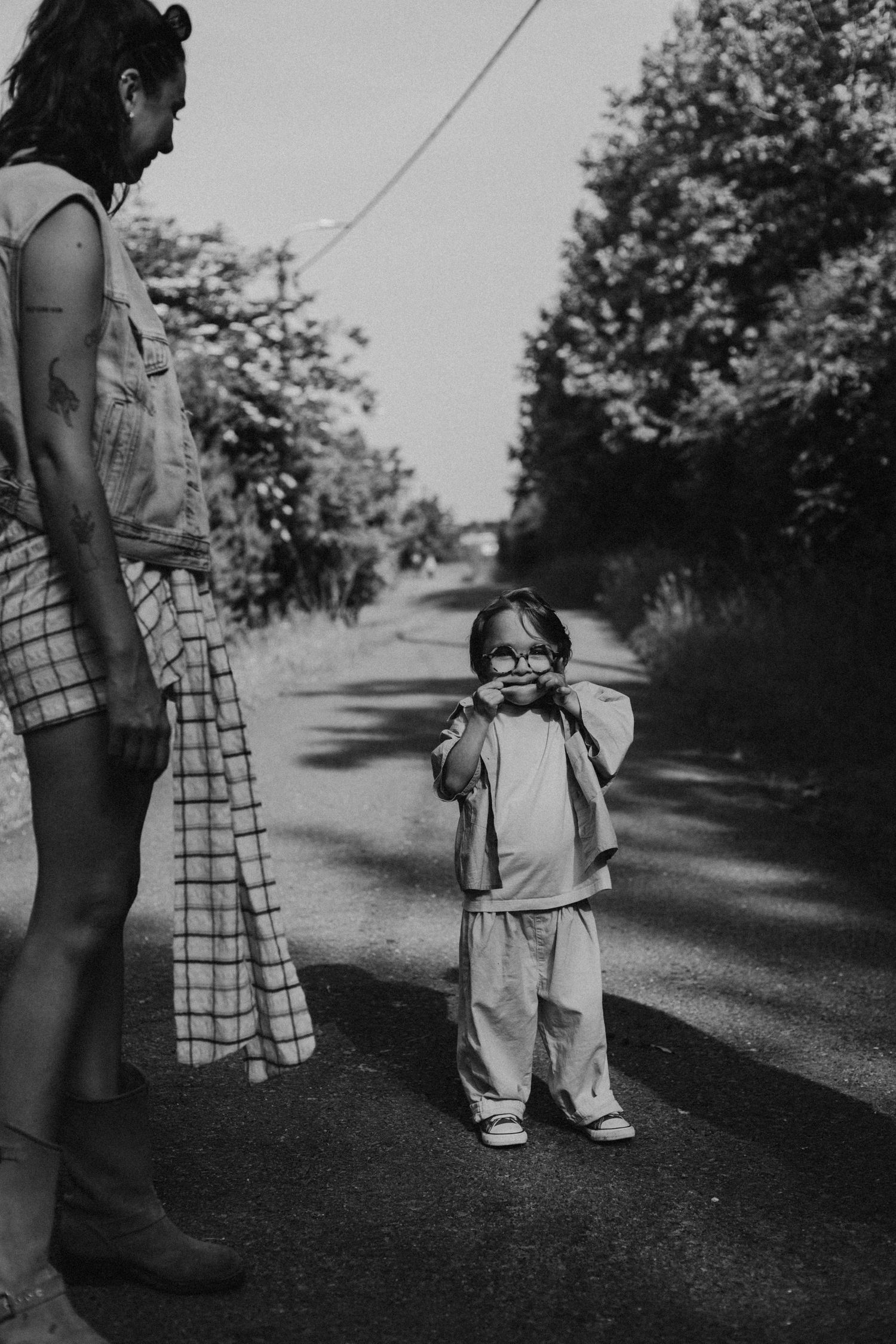Black and white photo of a toddler in glasses standing on a rural road beside an adult woman.