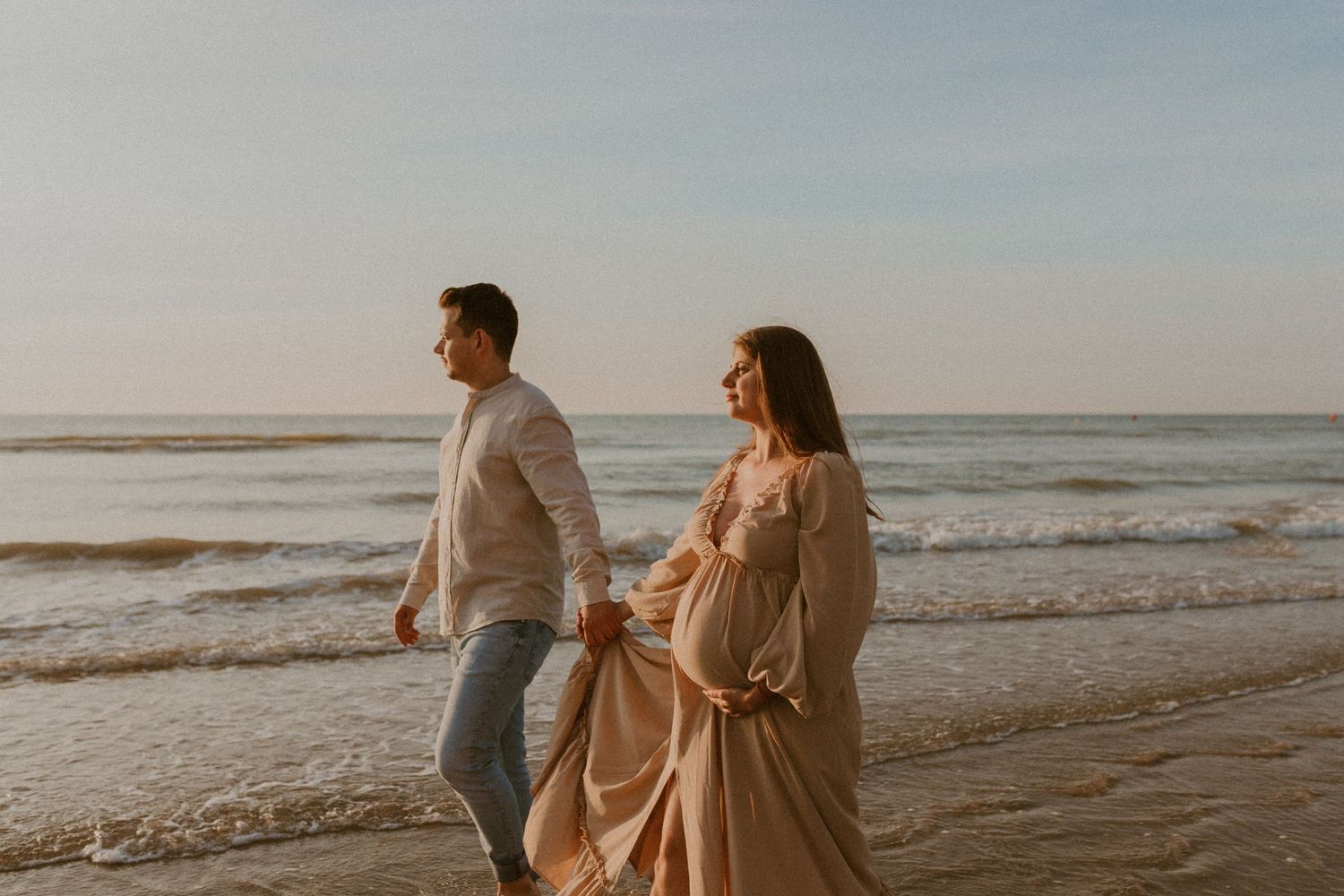 Couple in beige outfits walking along a misty beach shoreline at sunset.