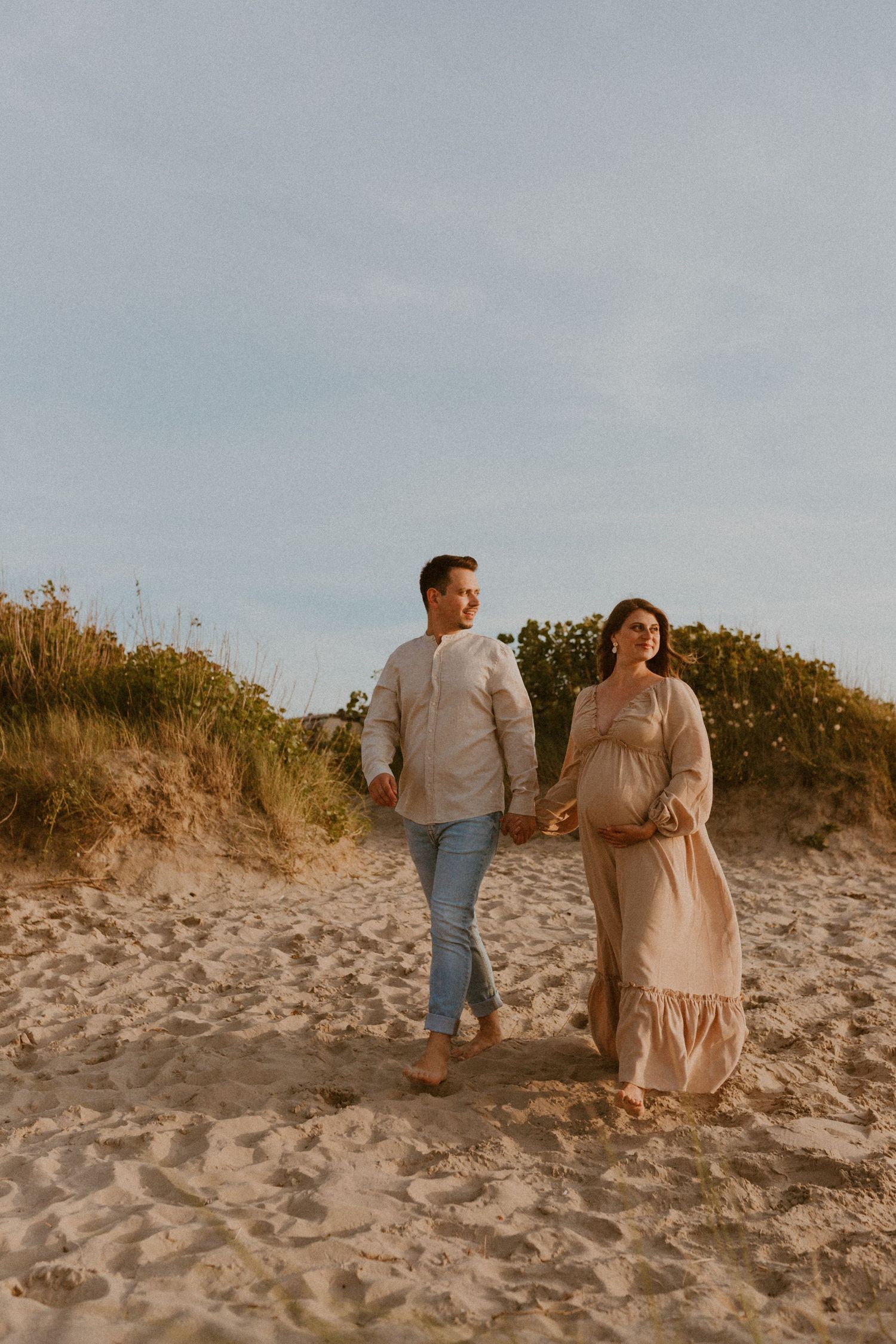 A couple walks together on a sandy beach at sunset with autumn foliage in the background.