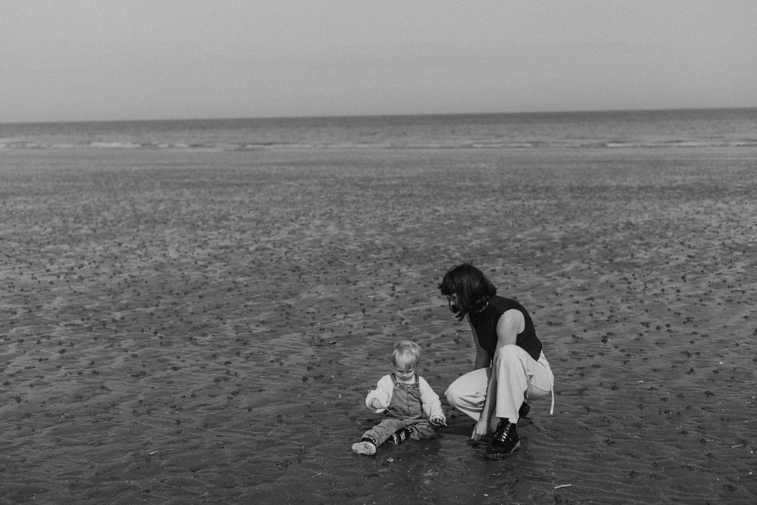 Black and white photo of a mother and baby playing together on a sandy beach by the ocean.