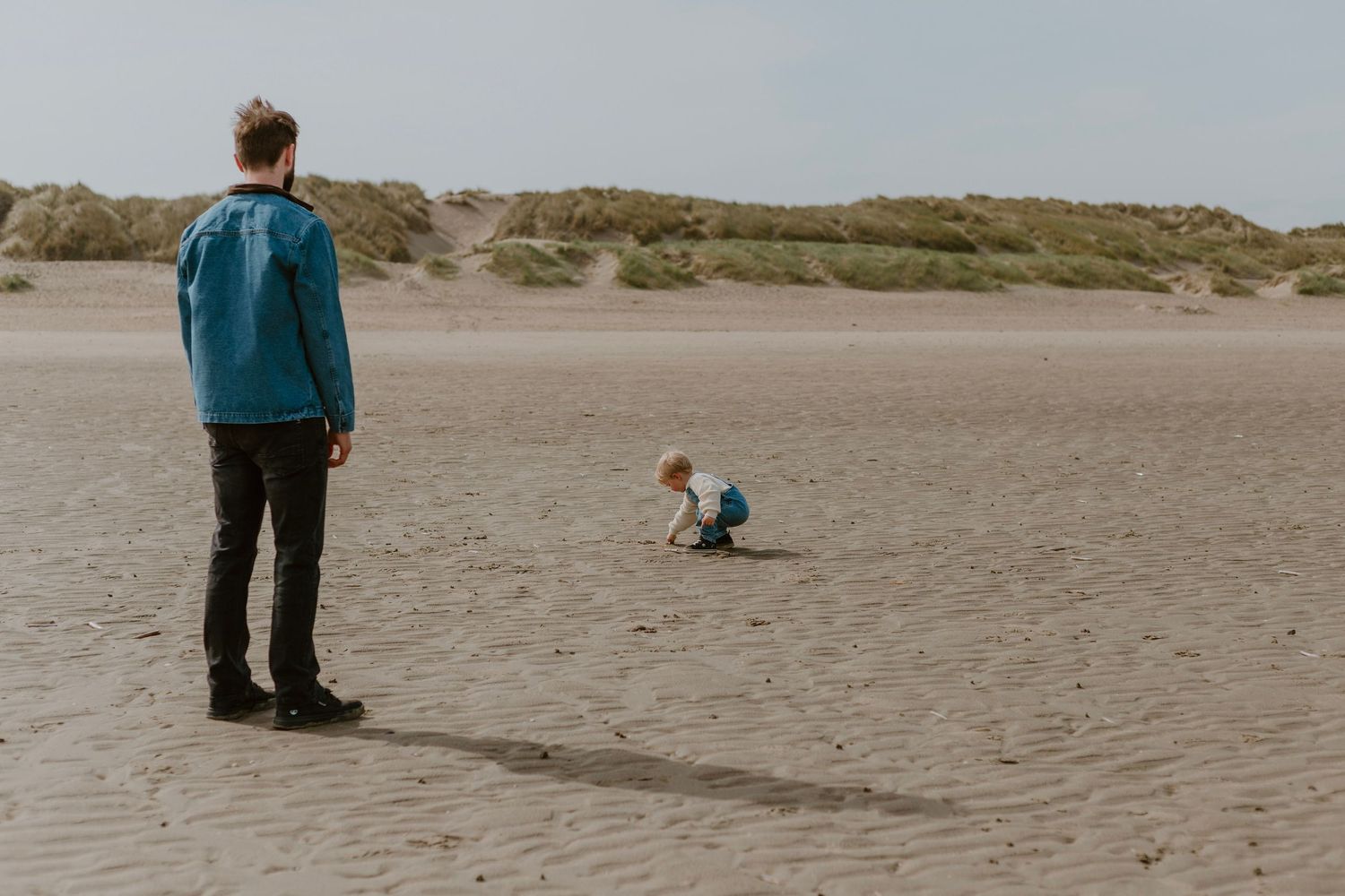 Adult watching a young child playing on a sandy beach with grass-covered dunes in the background.