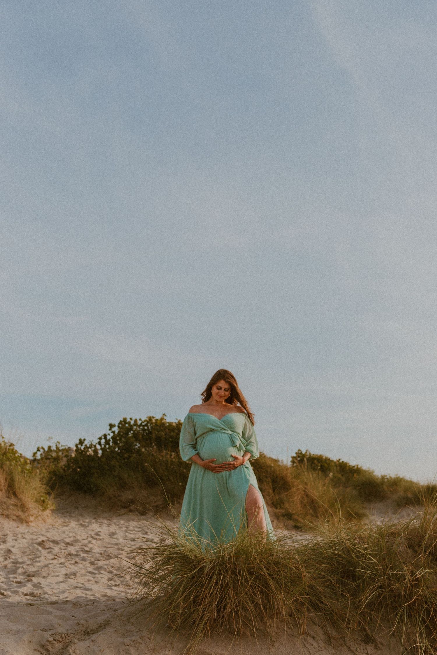 A woman in a flowing mint green dress poses gracefully among beach grass dunes at sunset.