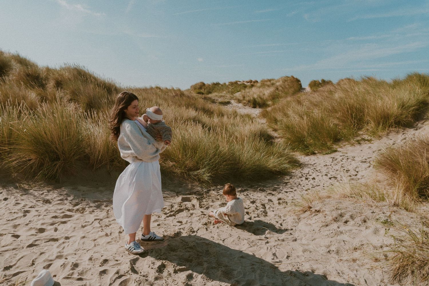 Two people in white outfits walk along a sandy beach path surrounded by tall grass on a sunny day.