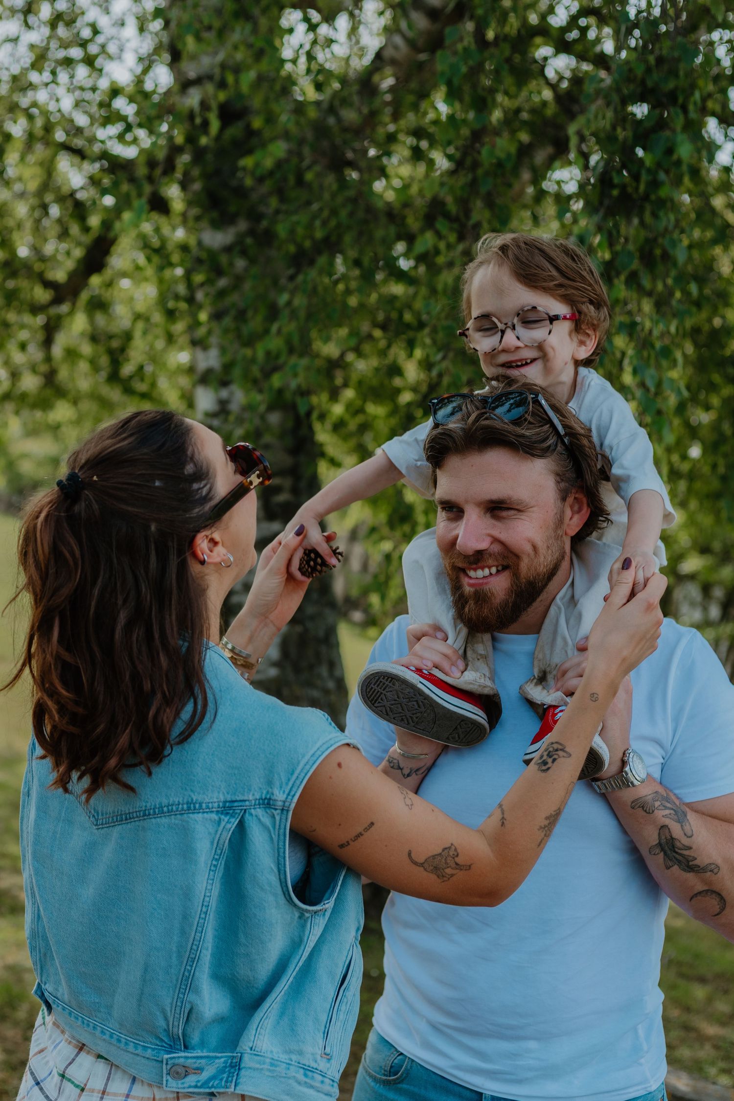 A tattooed couple smiles as their young child with glasses sits on dad's shoulders in a sunny park.