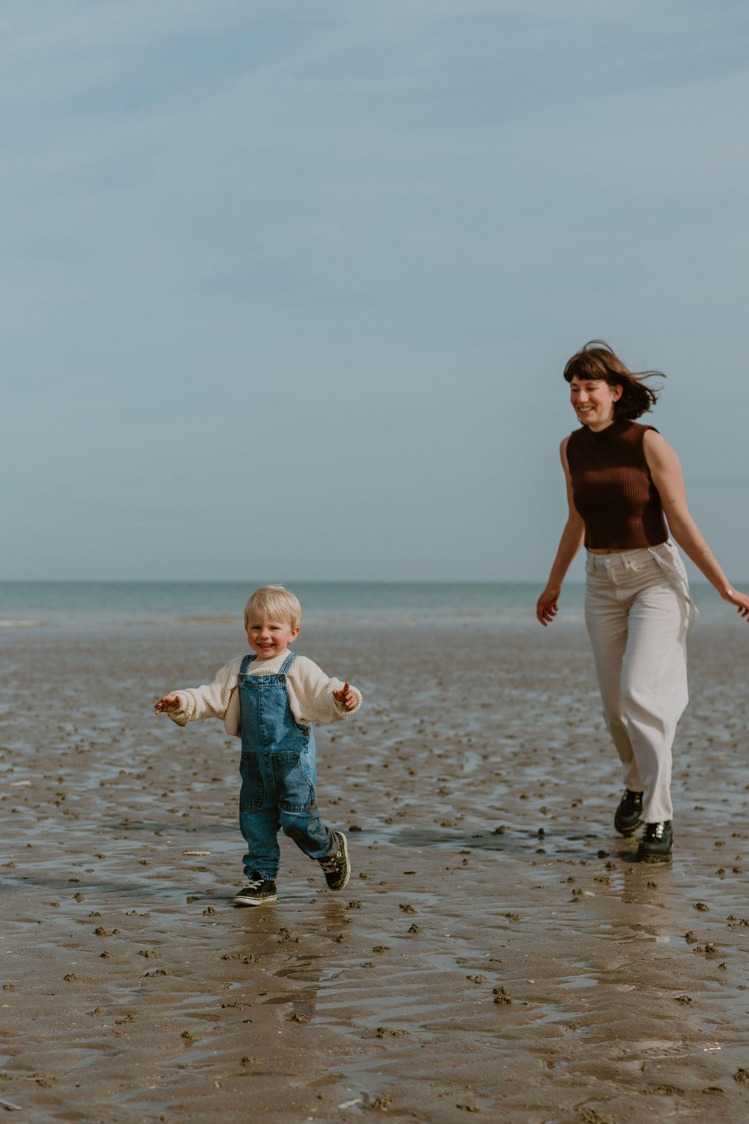 A woman and young child joyfully running on a wet sandy beach with calm ocean waves in the background.