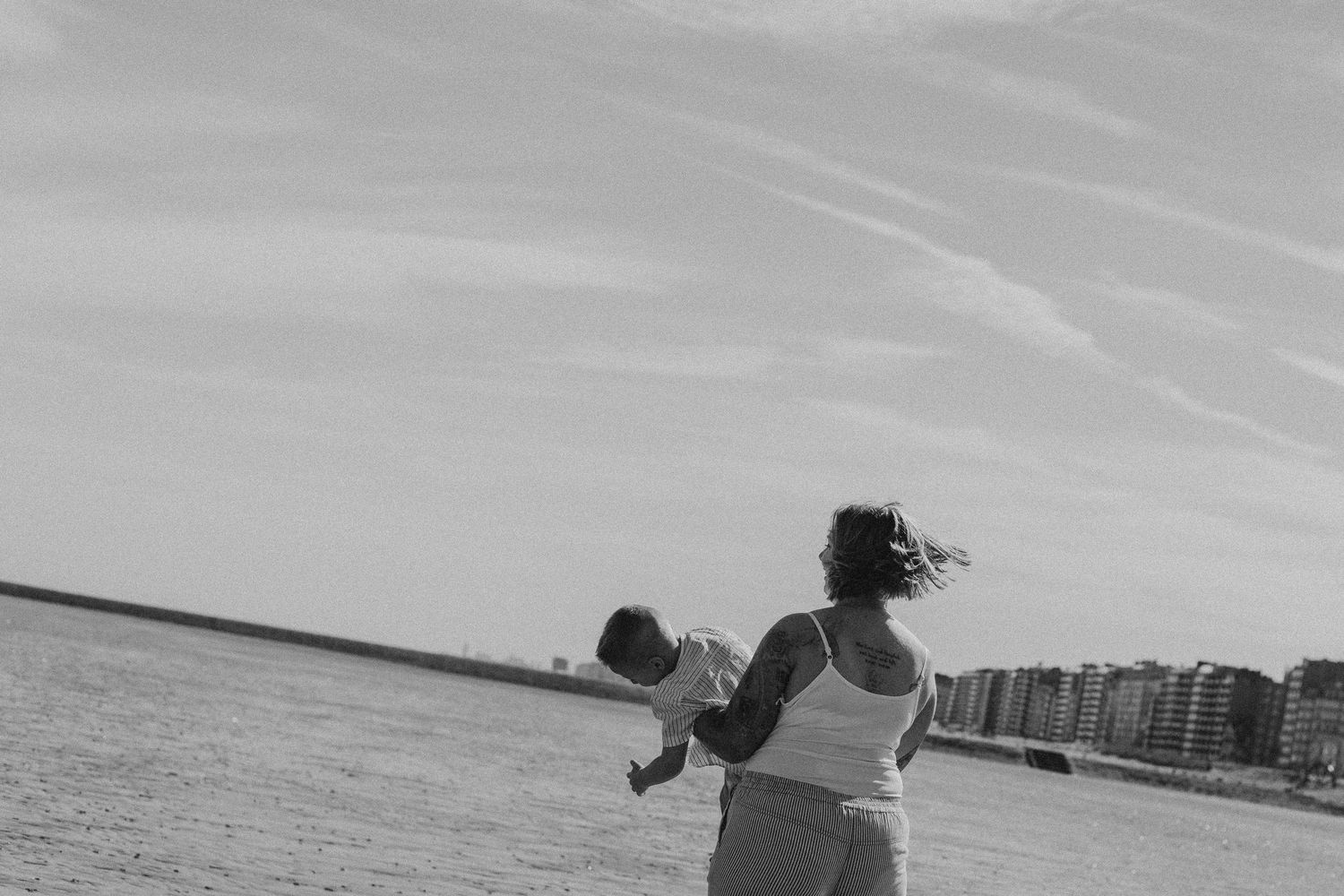 Black and white photo of a woman holding a baby on a beach with buildings in the background.