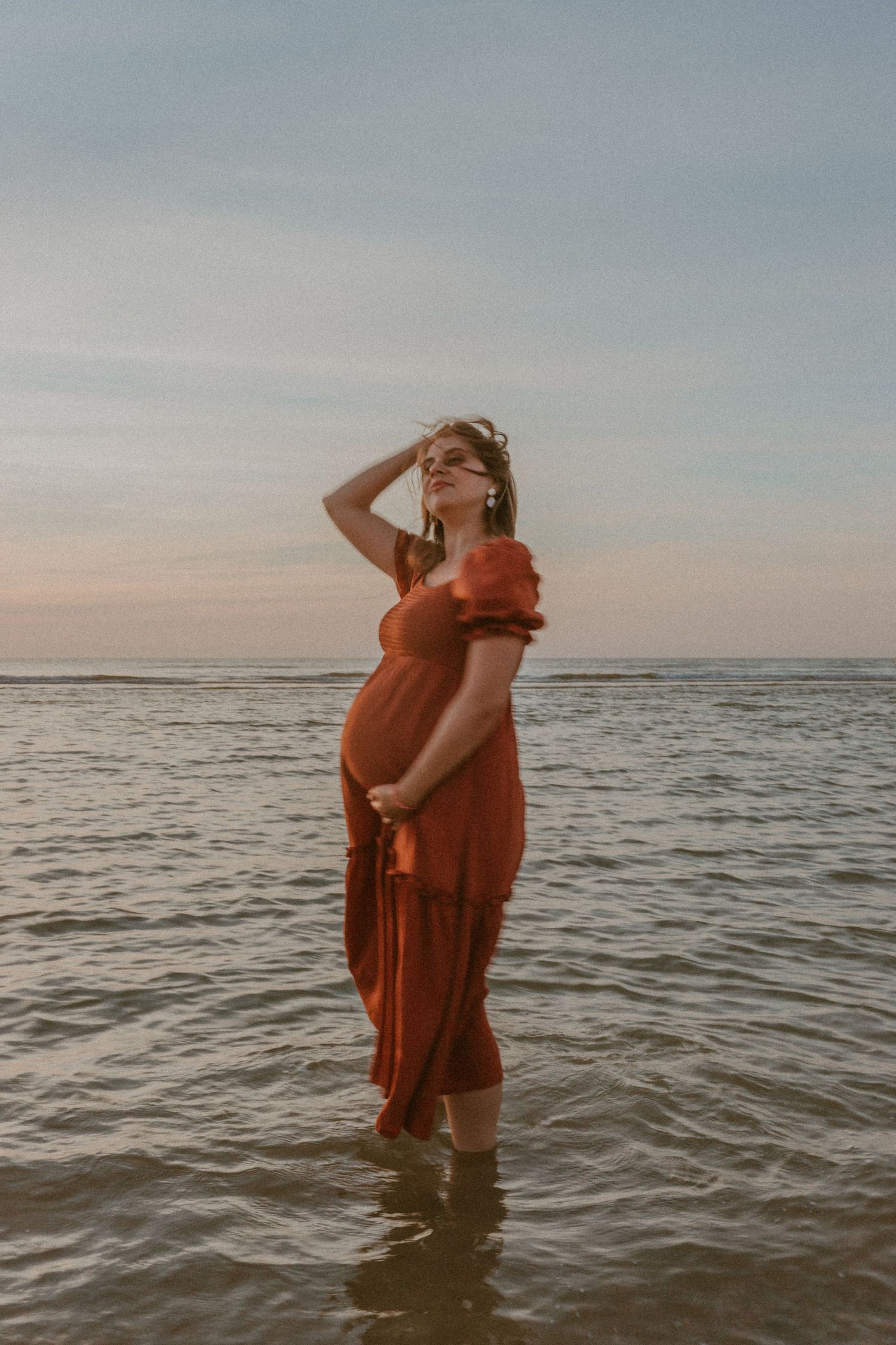 A pregnant woman in a flowing red dress stands in shallow ocean water at sunset, hand raised to her head in a dreamy pose.