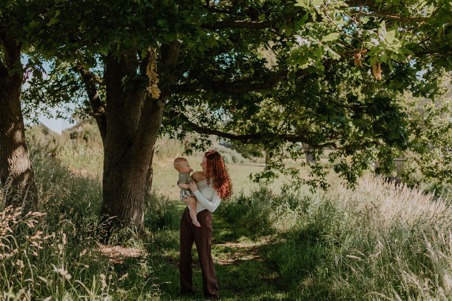 Couple embracing under a large green tree in a sunlit meadow, romantic outdoor portrait photography.