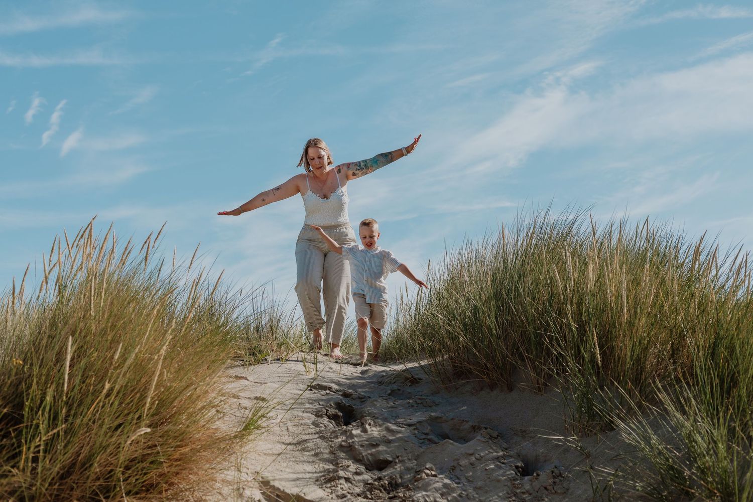 A joyful woman and child run through sandy beach dunes with tall grass under a bright blue sky.