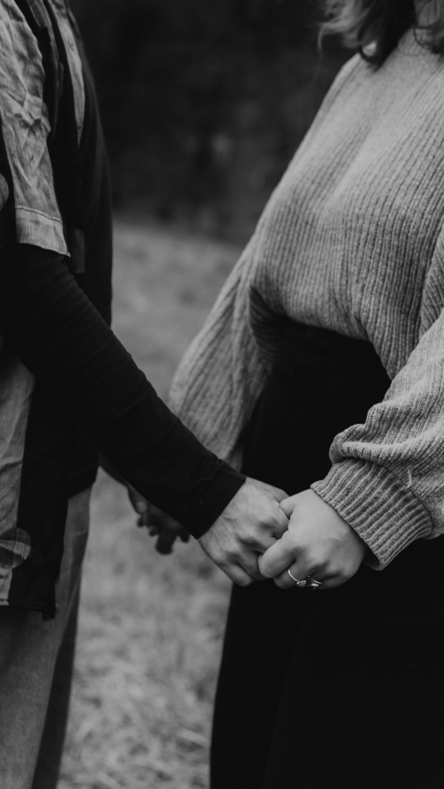 A close-up black and white photo of two people holding hands while wearing sweaters and dark clothing.