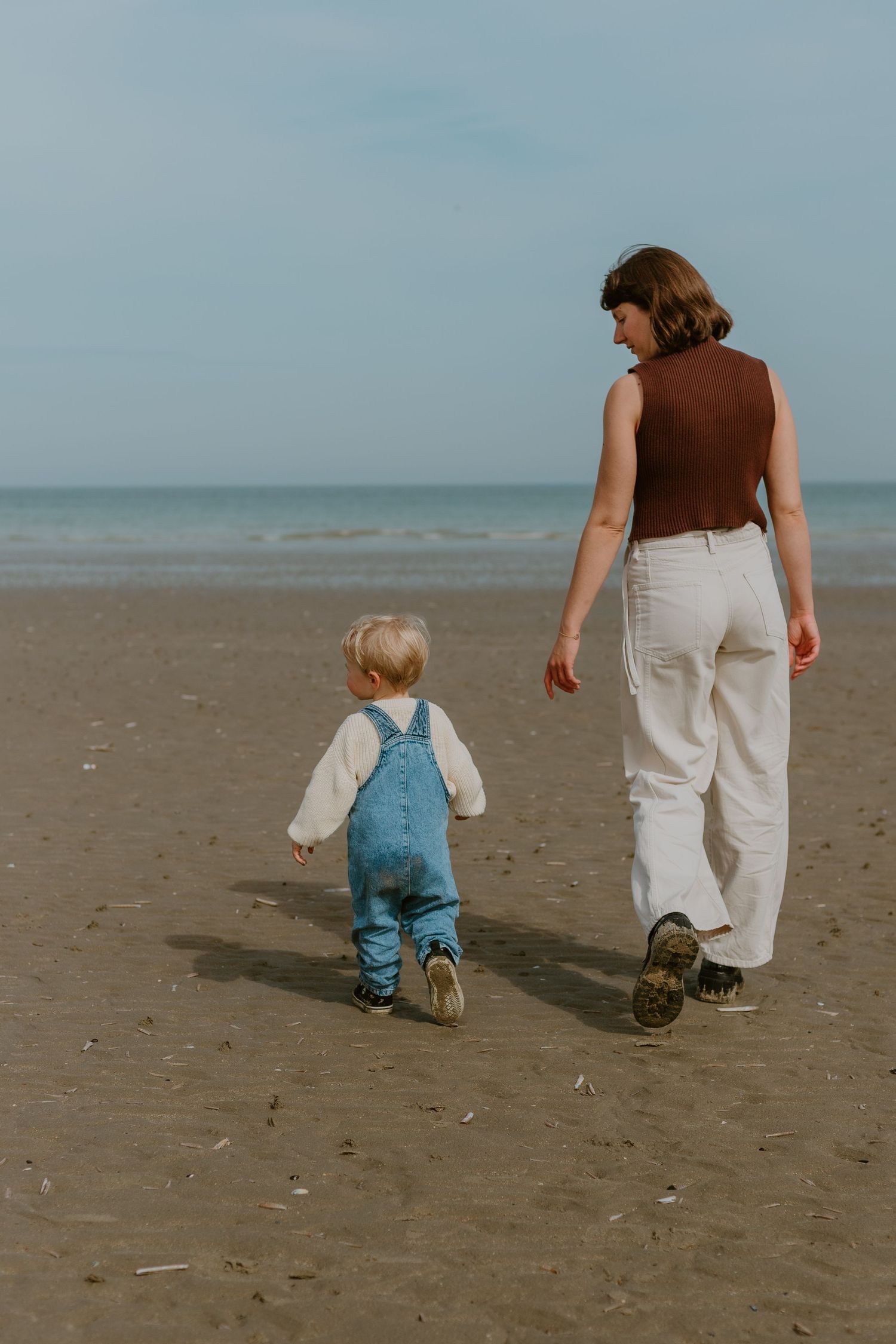 A mother and toddler walking hand in hand on a sandy beach toward the ocean on a clear day.