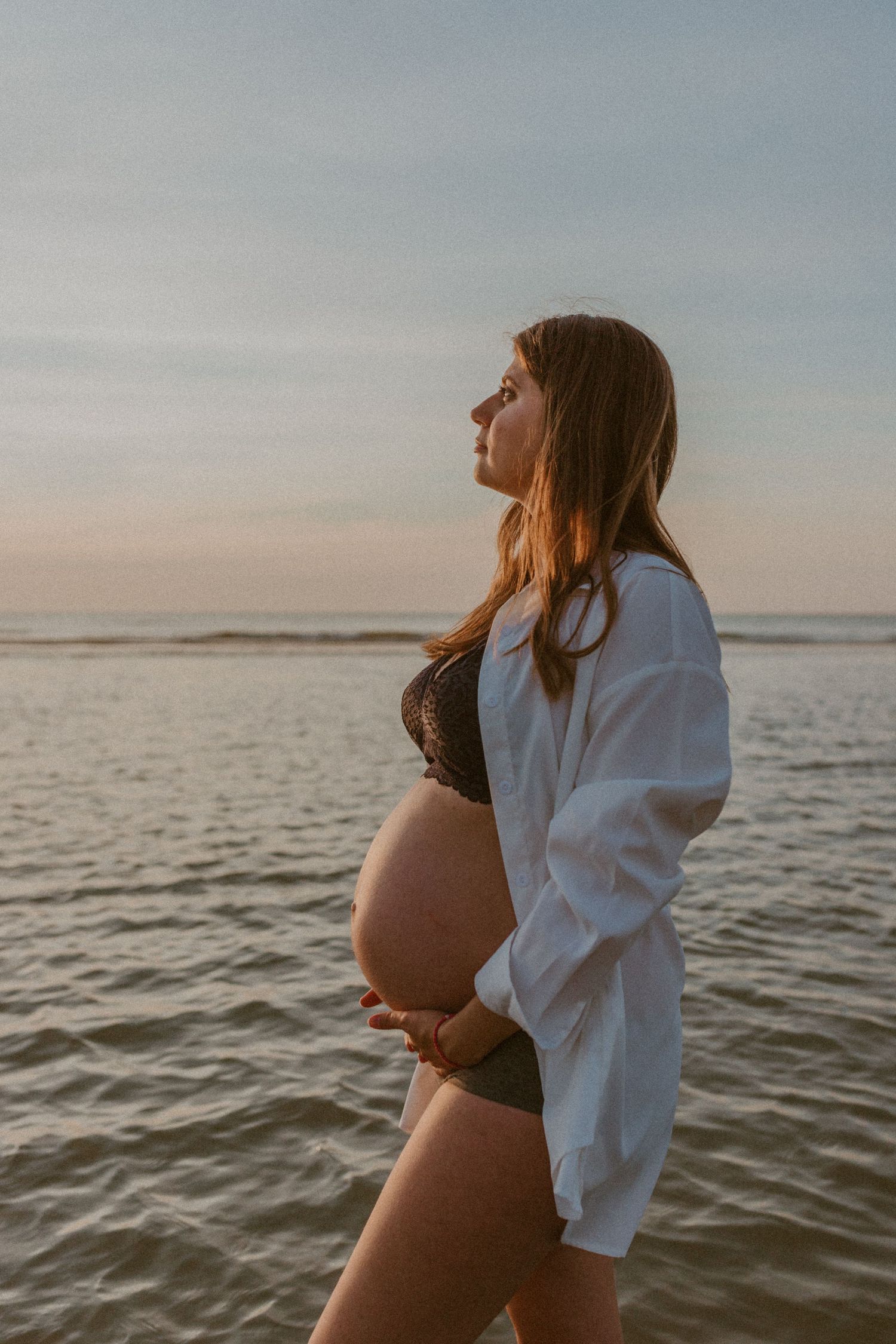 Silhouetted pregnant woman in black swimwear and white cover-up stands at ocean's edge during golden sunset.