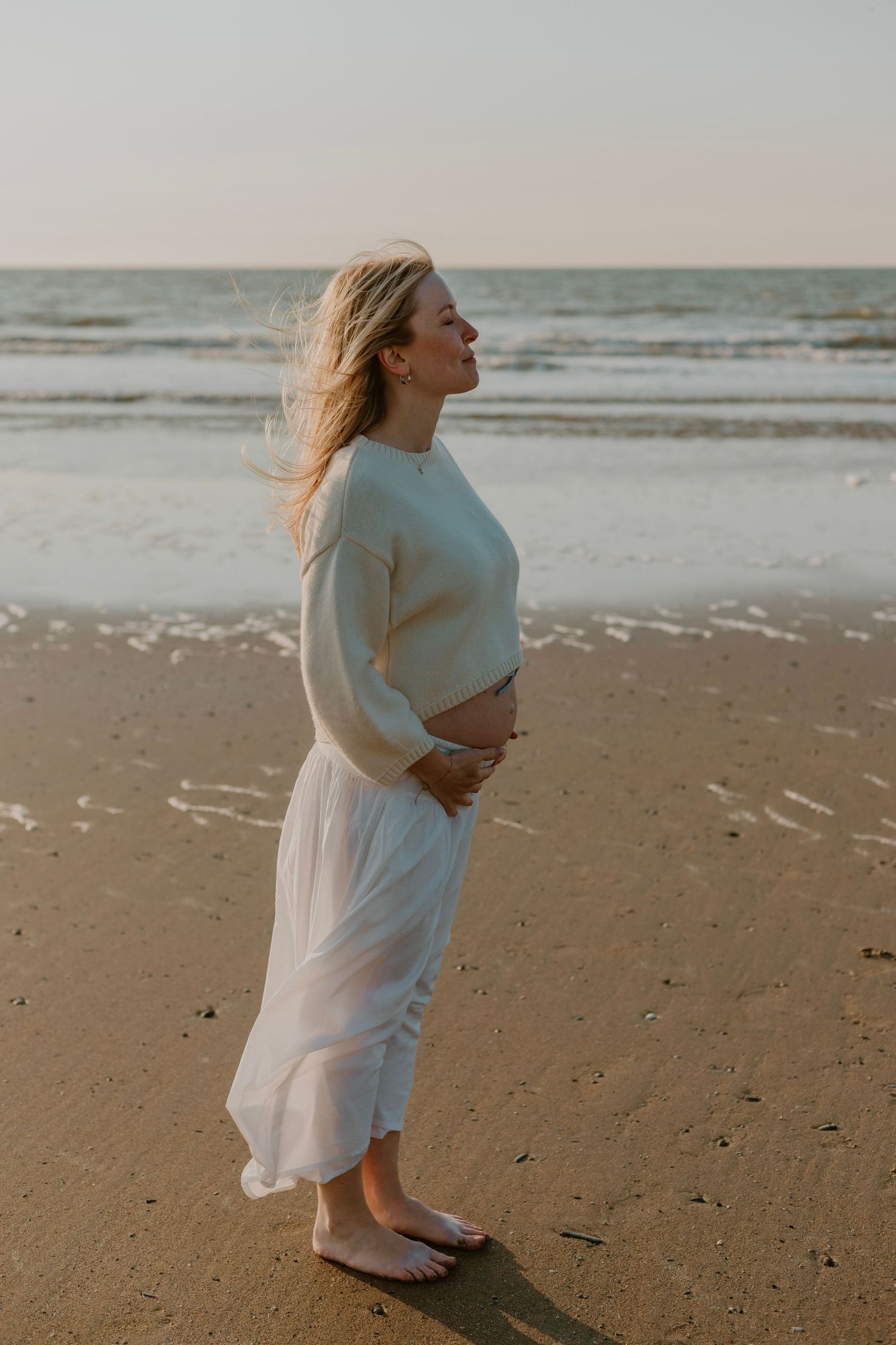 Pregnant woman in white skirt and sweater standing barefoot on sandy beach at sunset, cradling her baby bump.