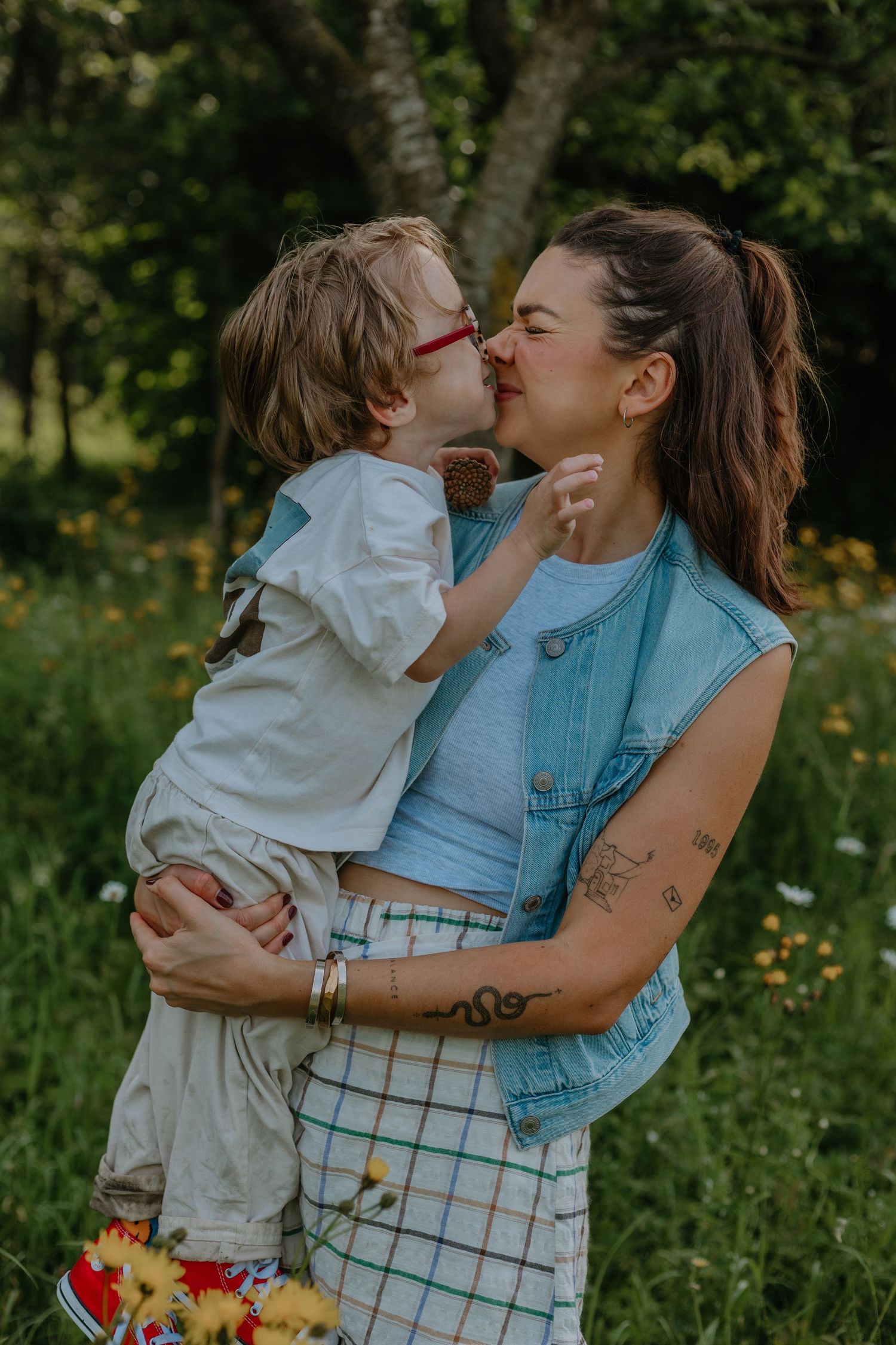 A mother holds and kisses her young child in a lush green meadow, both sharing a tender moment outdoors.
