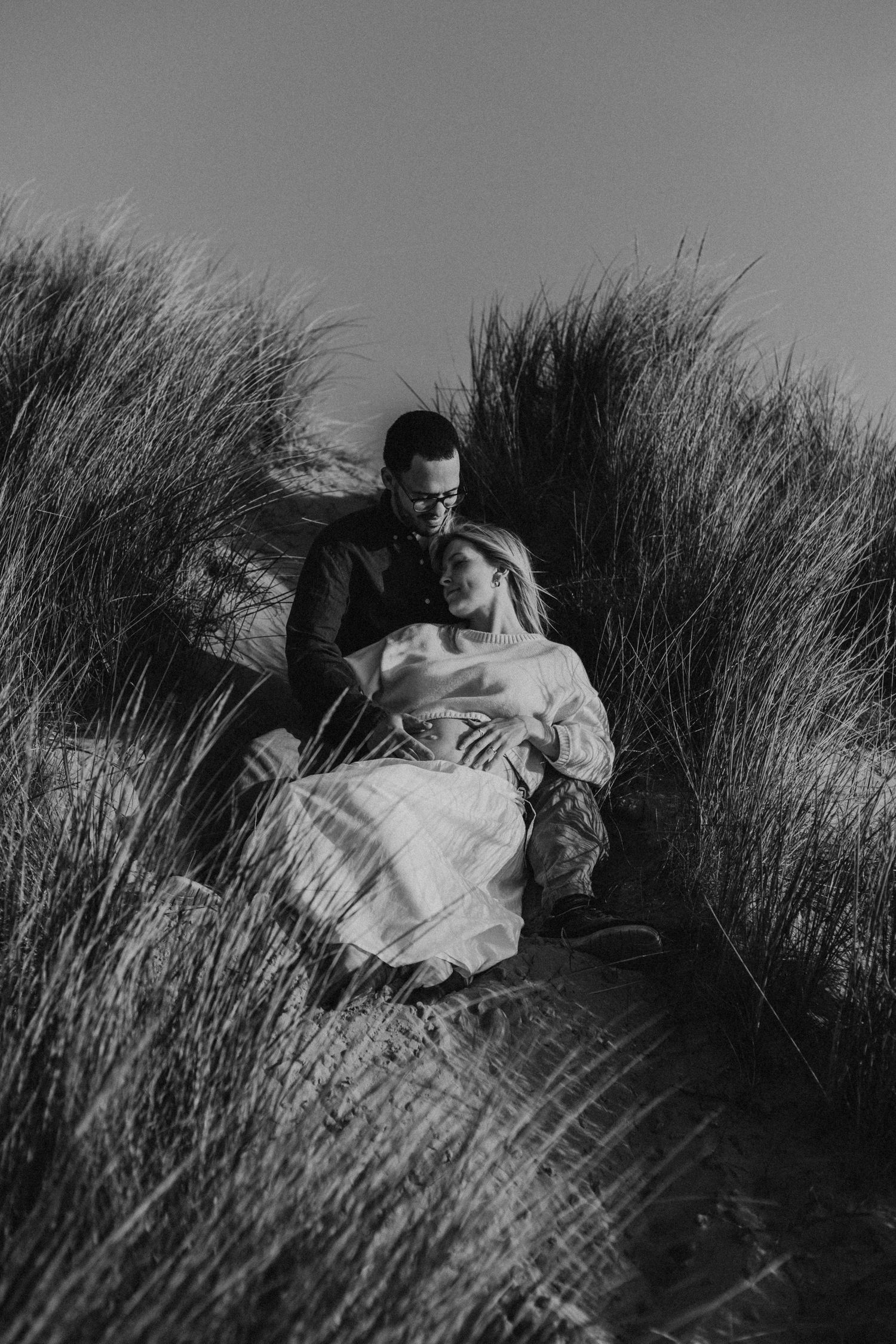 Black and white photo of a couple sitting together in tall beach grass, sharing an intimate moment.