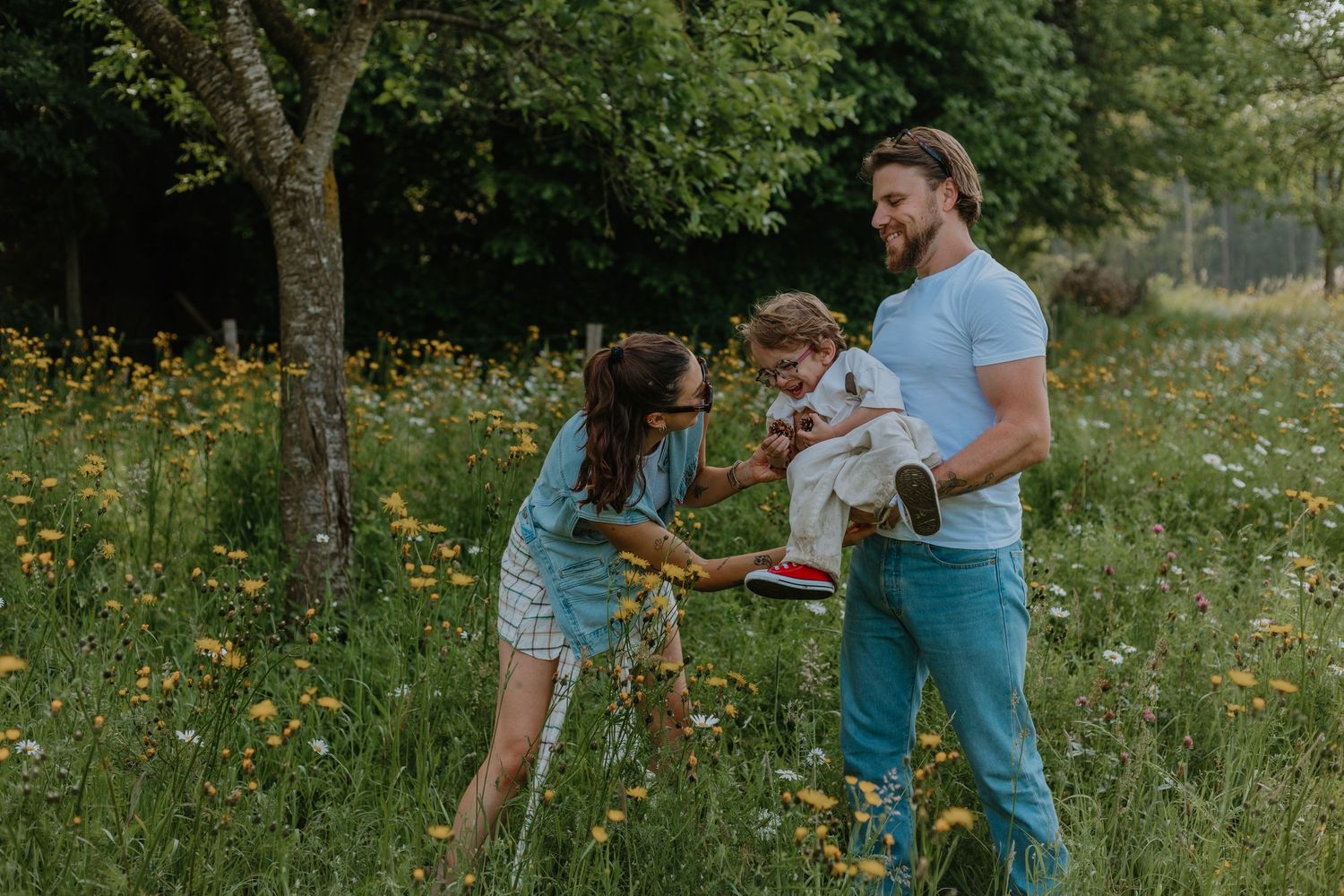 Family playing outdoors in a wildflower meadow, parents lifting toddler with joy on a sunny summer day.