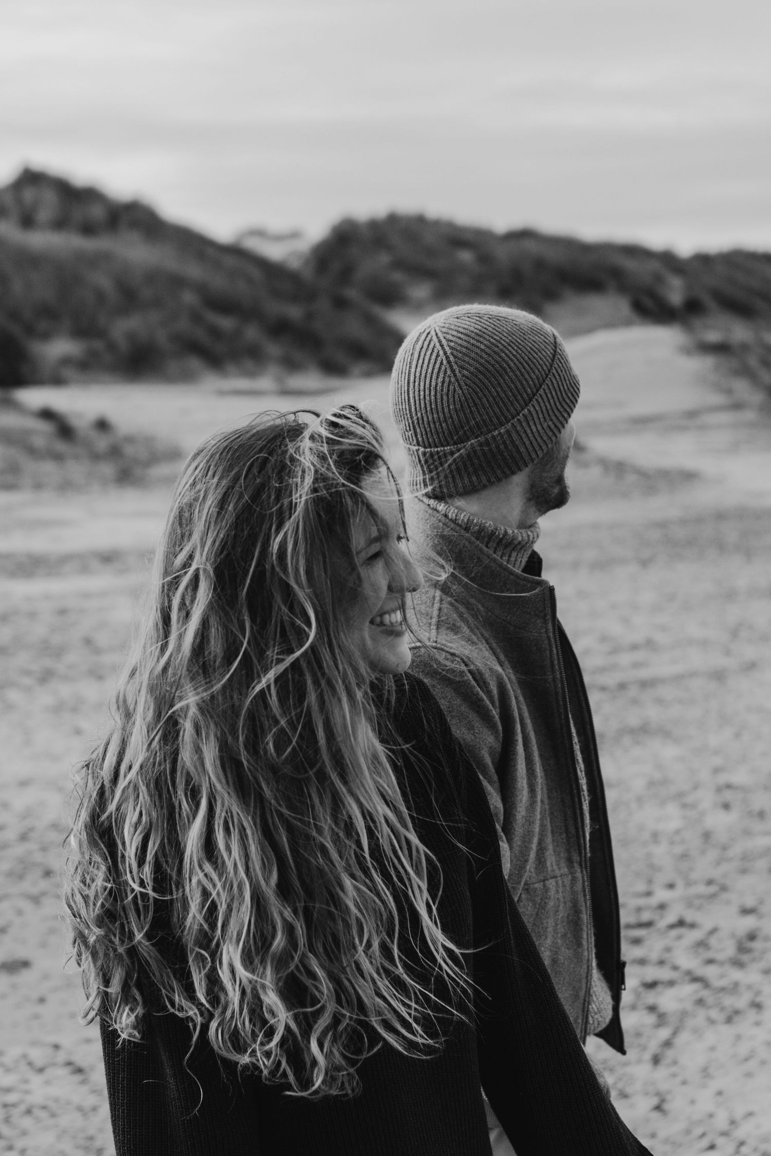 Black and white photo of a couple walking on a beach, one wearing a beanie hat and one with long wavy hair.