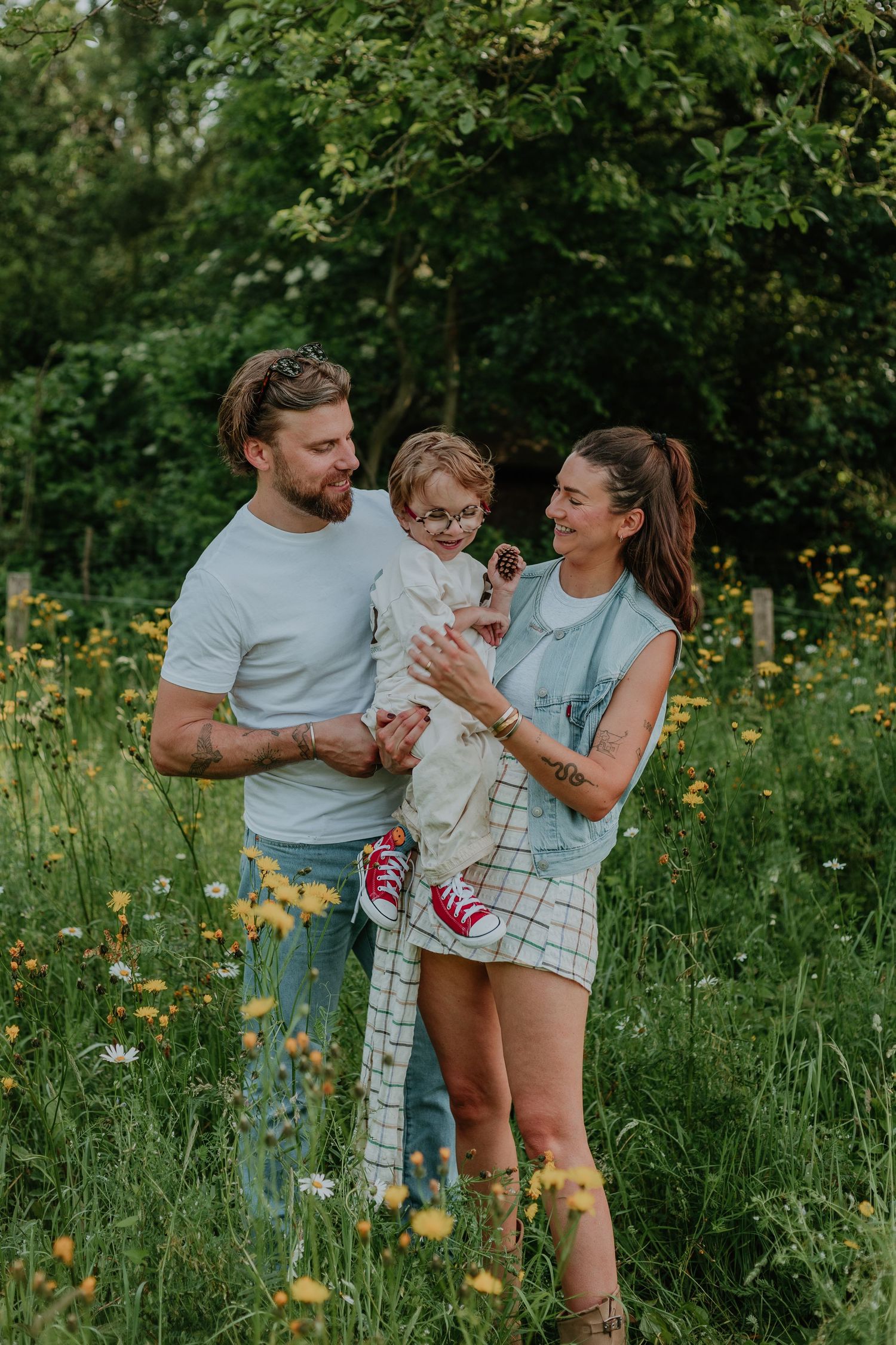 A young family with a toddler in red sneakers poses in a sunny wildflower meadow surrounded by lush green trees.