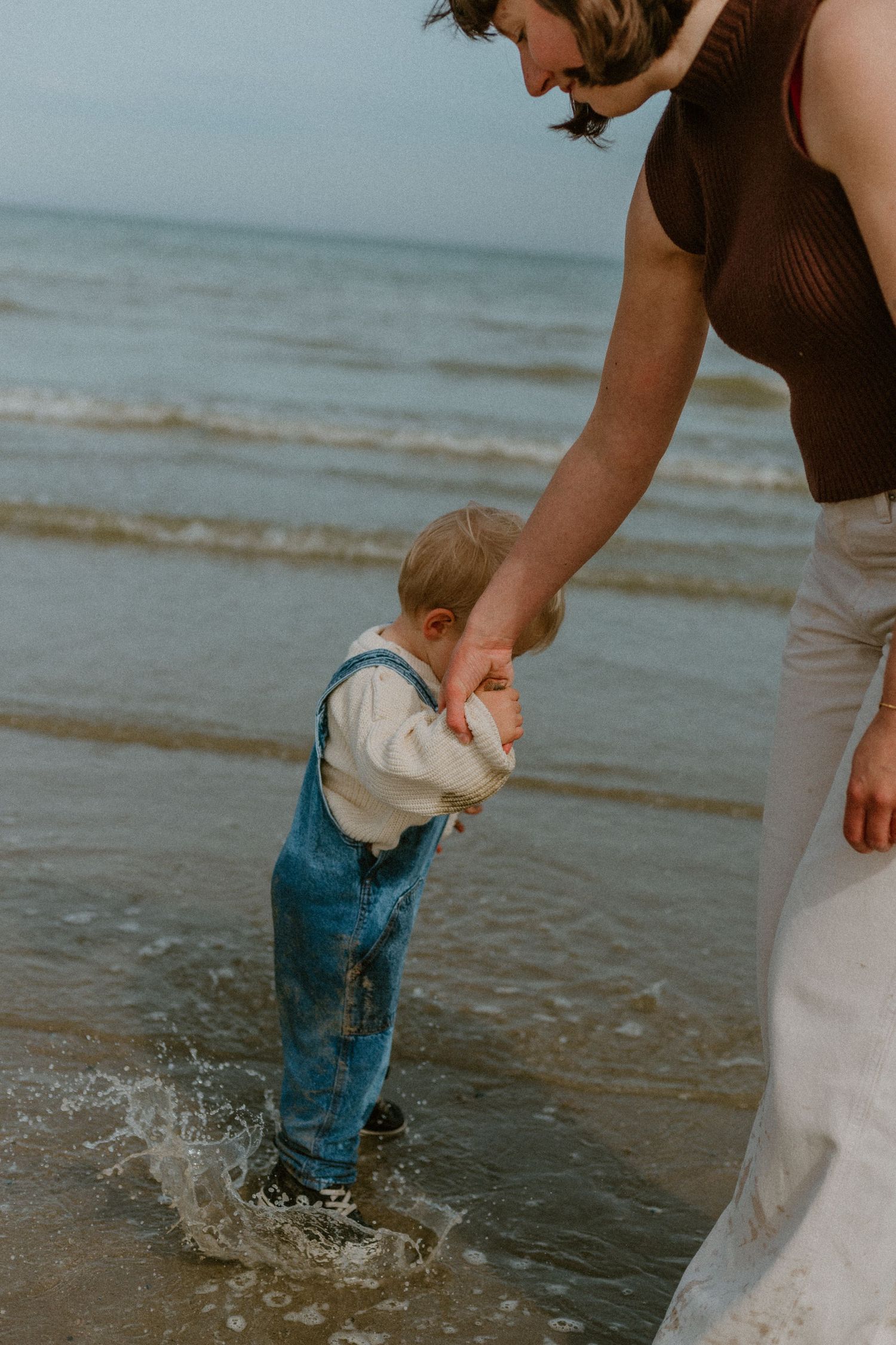 Adult holding toddler's hands while child splashes in ocean waves on sandy beach, wearing denim overalls.