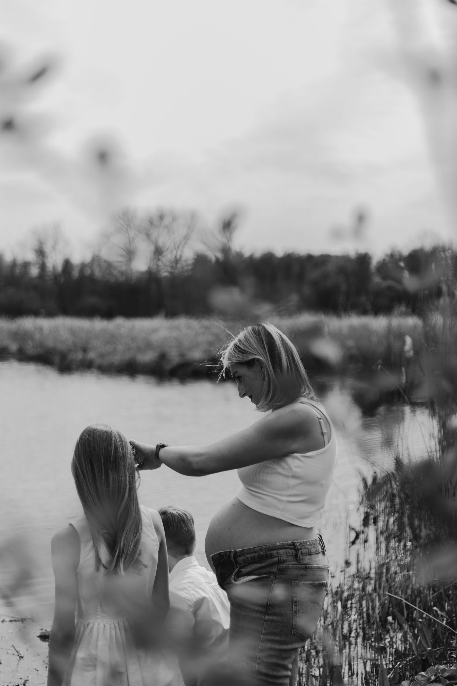 Black and white photo of a pregnant woman tenderly styling her daughter's hair near a serene lake outdoors.