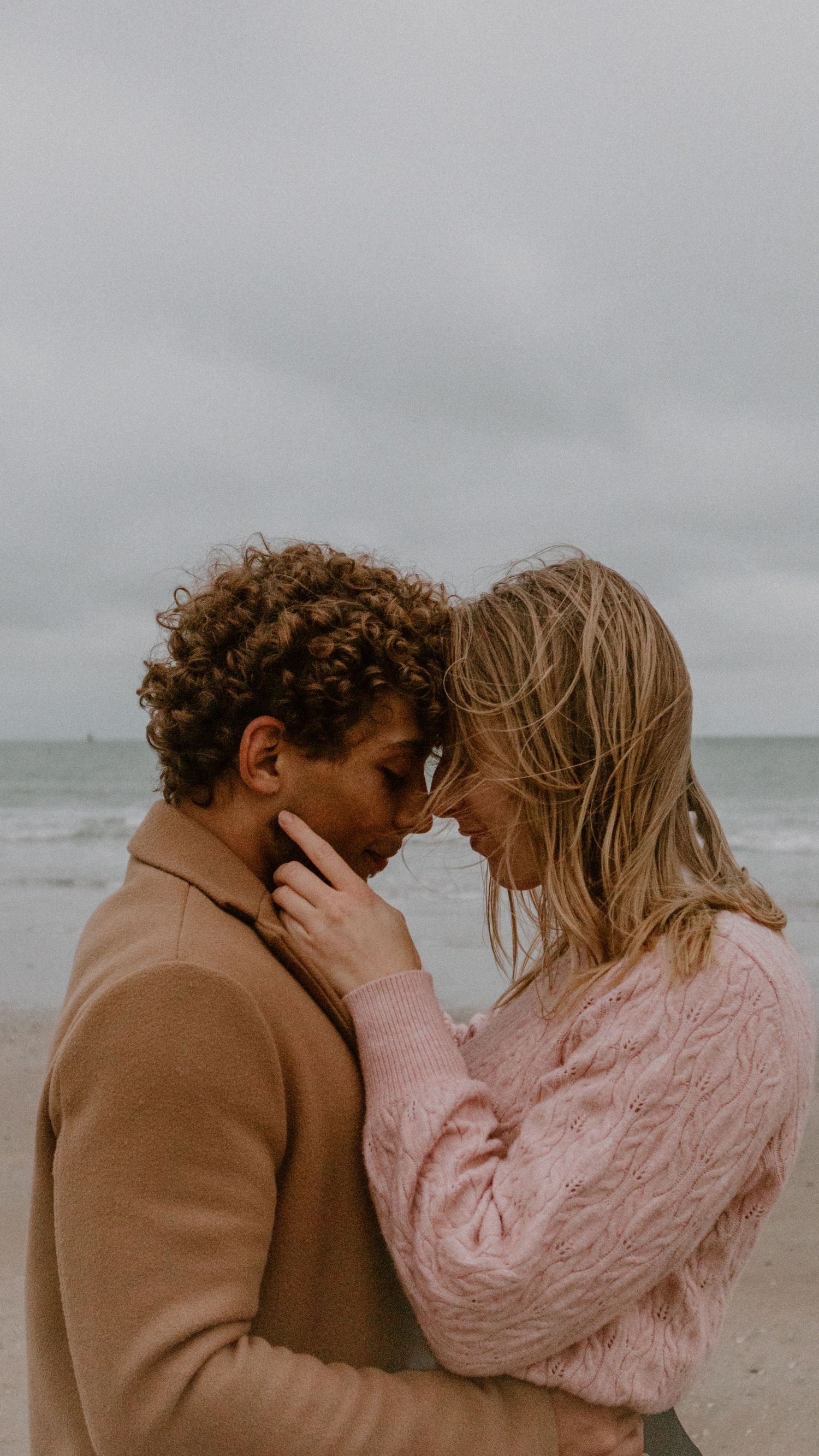 A couple shares an intimate moment on a cloudy beach, with one wearing a brown coat and the other in pink sweater.