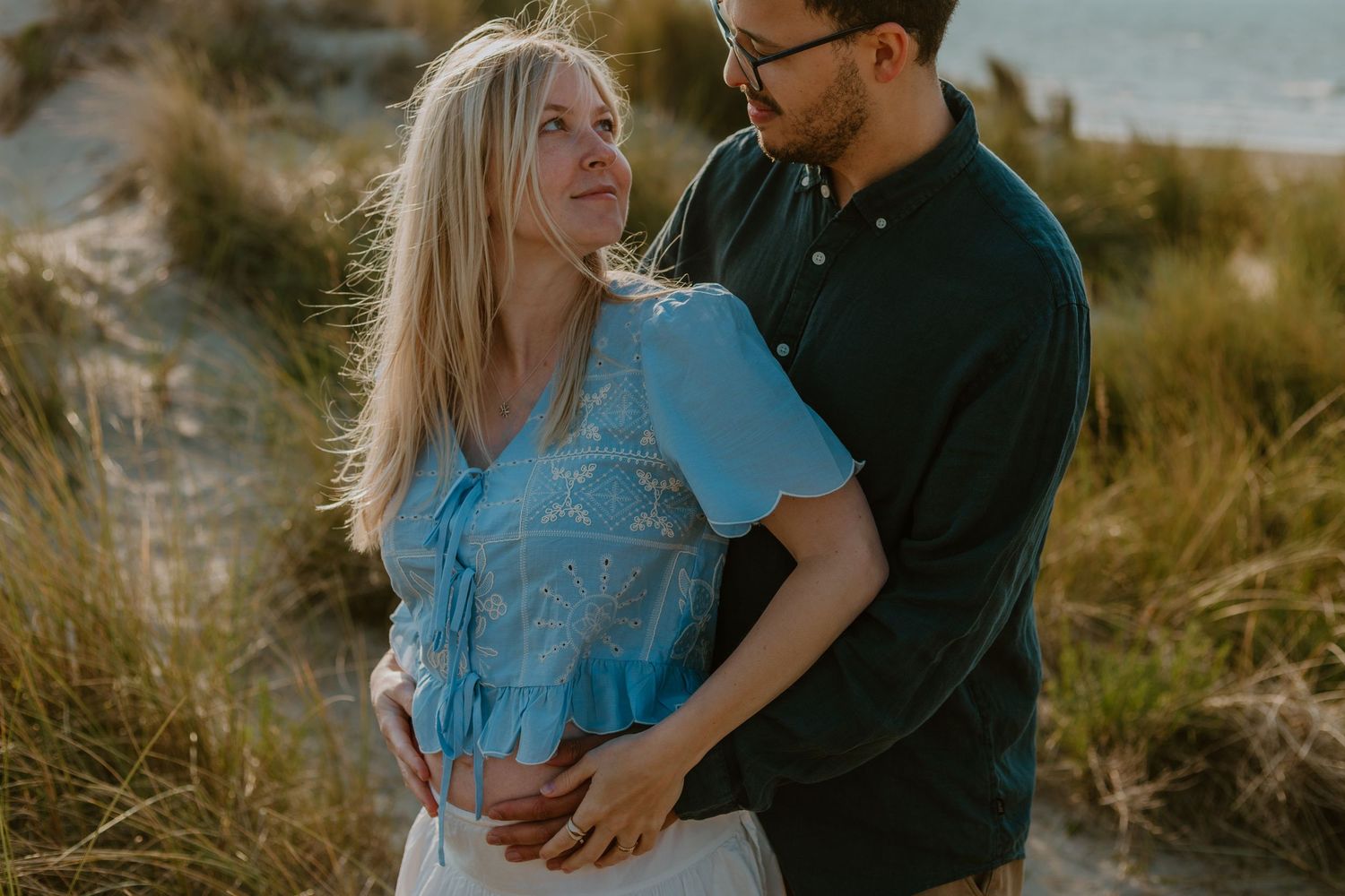 A couple embraces tenderly on a sandy beach, the woman in a blue top and the man in a dark green shirt.