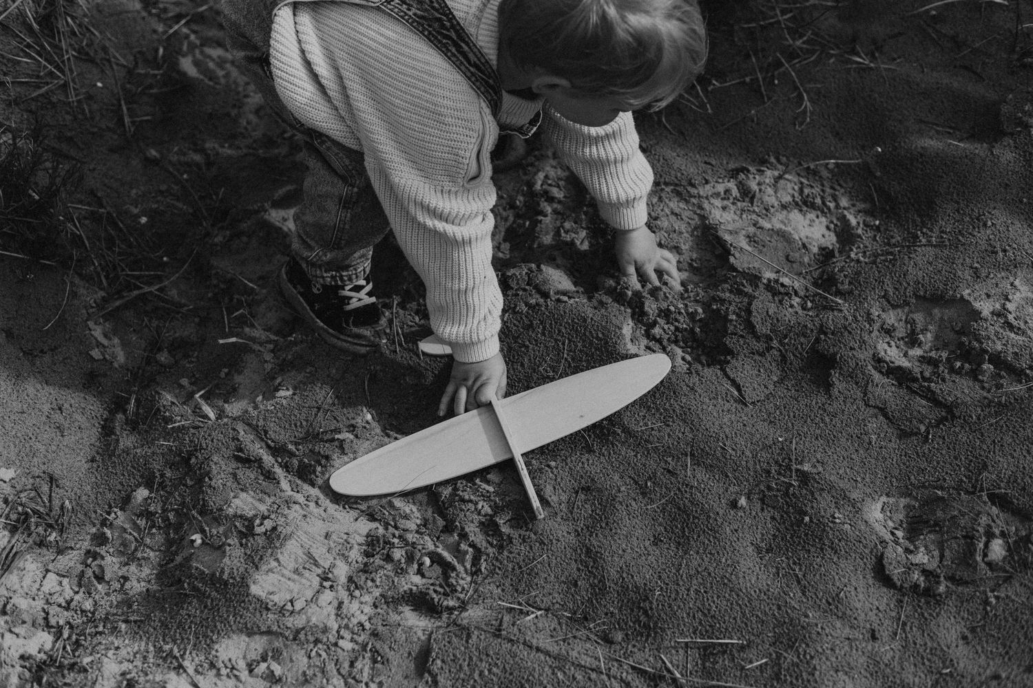 Black and white photo of a child playing with a toy glider plane in sandy dirt outdoors.