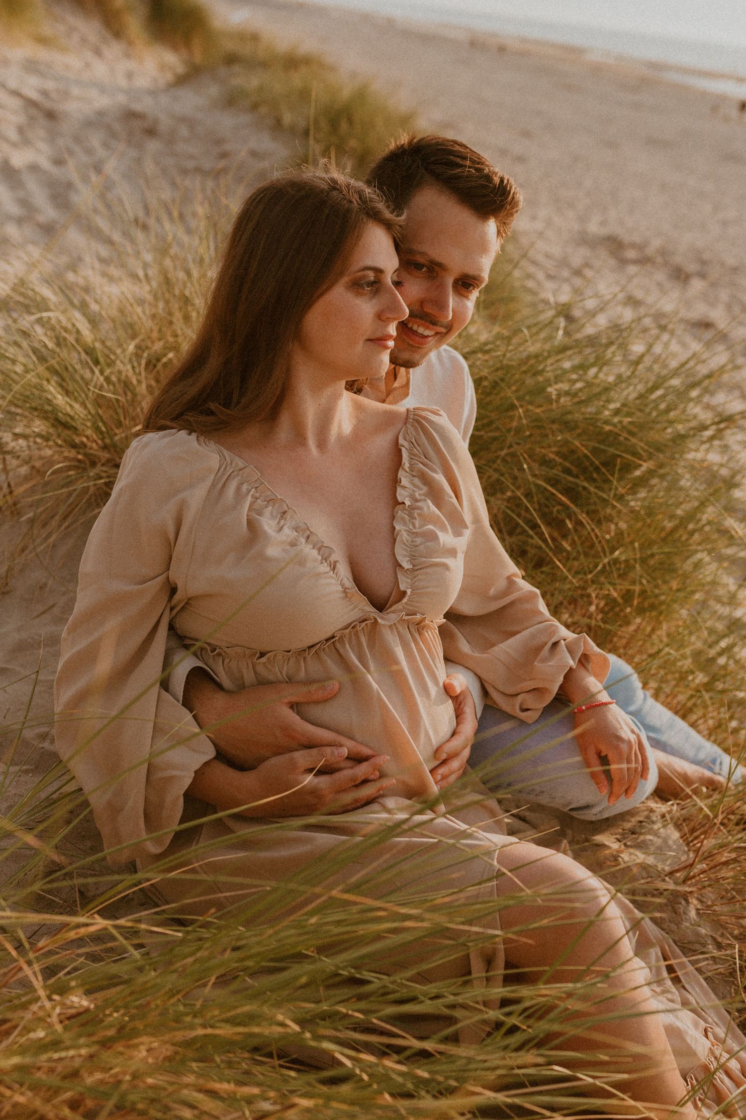 Romantic couple embraces in tall beachside grass during golden hour sunset photoshoot.