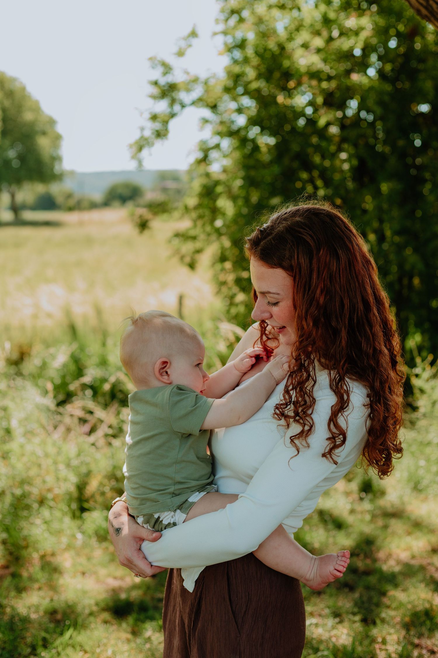 A red-haired woman holds her baby outdoors in a sunny green field, as the child playfully touches her face.