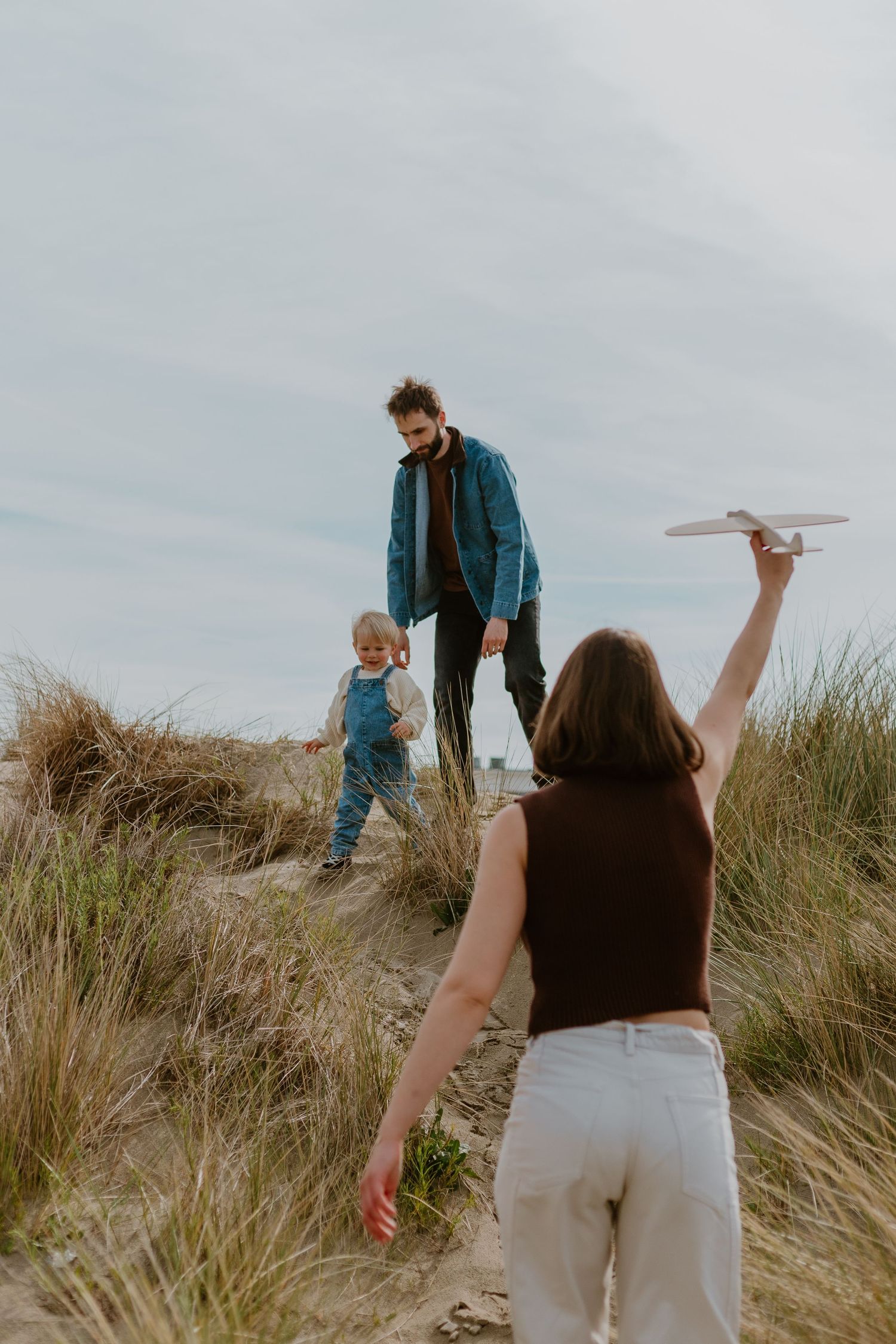 Family playing on sand dunes, woman holding paper airplane toward man and toddler on grassy beach.