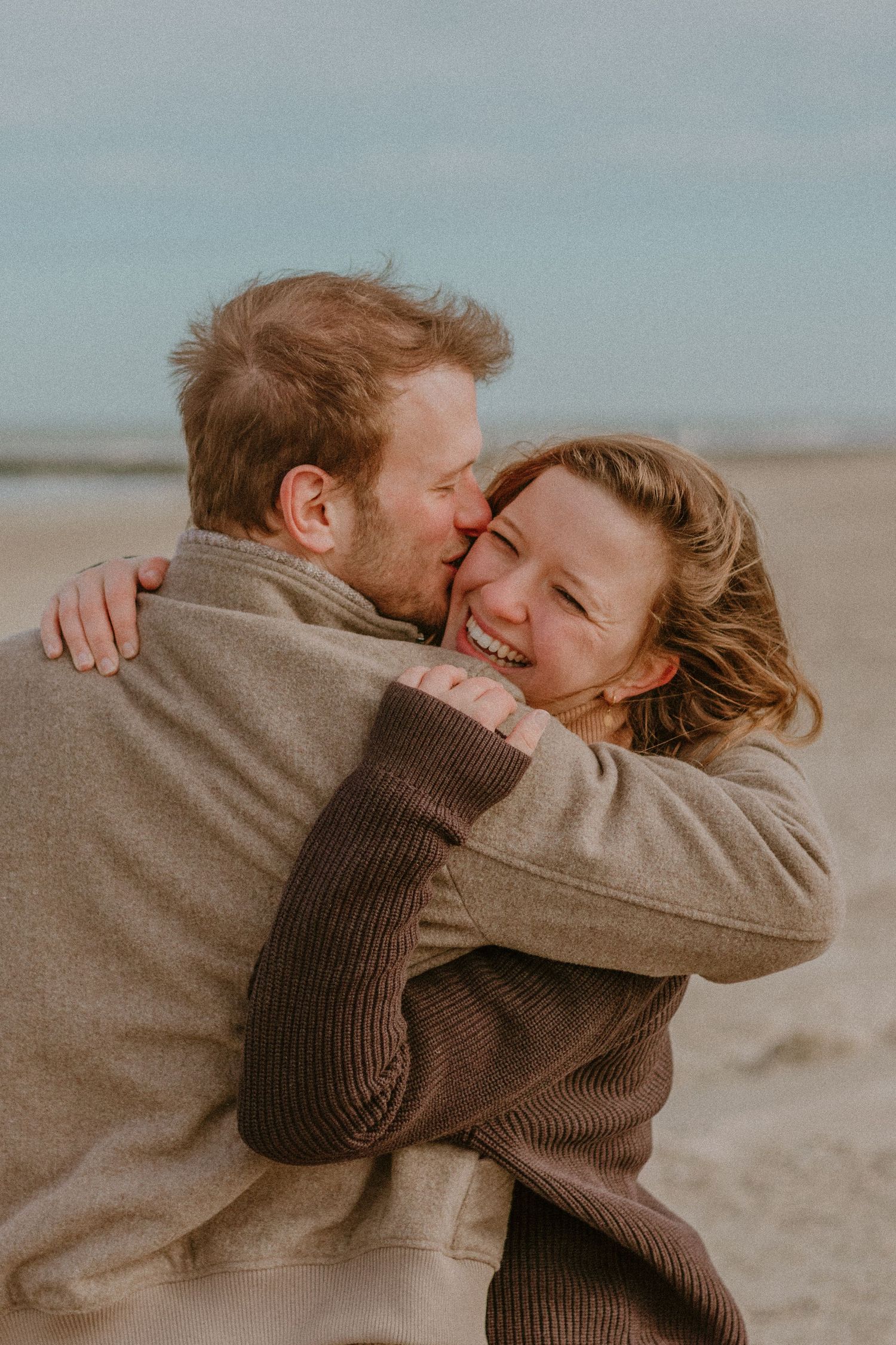 A couple in warm sweaters embrace and laugh together on a cloudy beach.