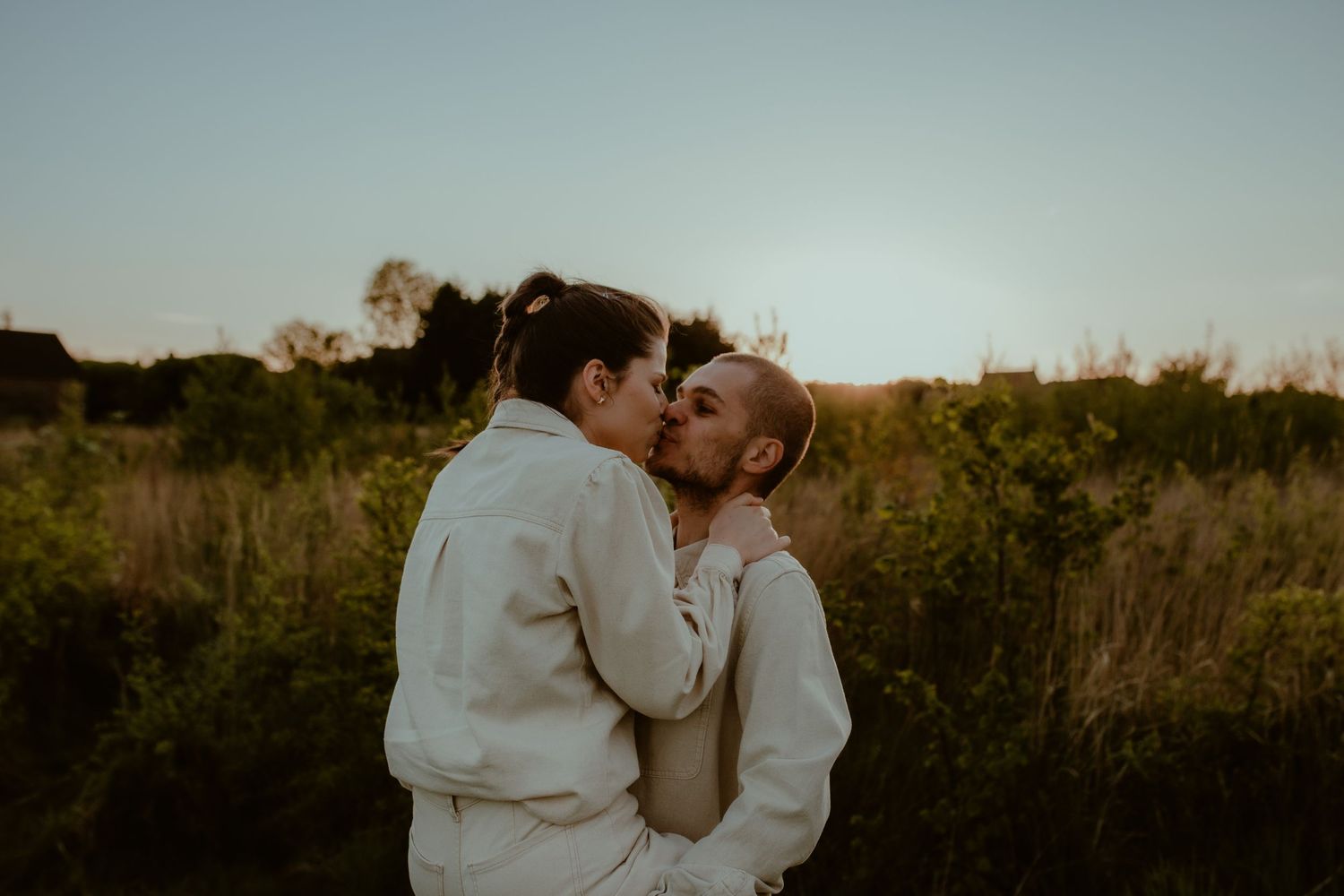 Two people in matching cream sweaters share a tender moment in a golden field at sunset.