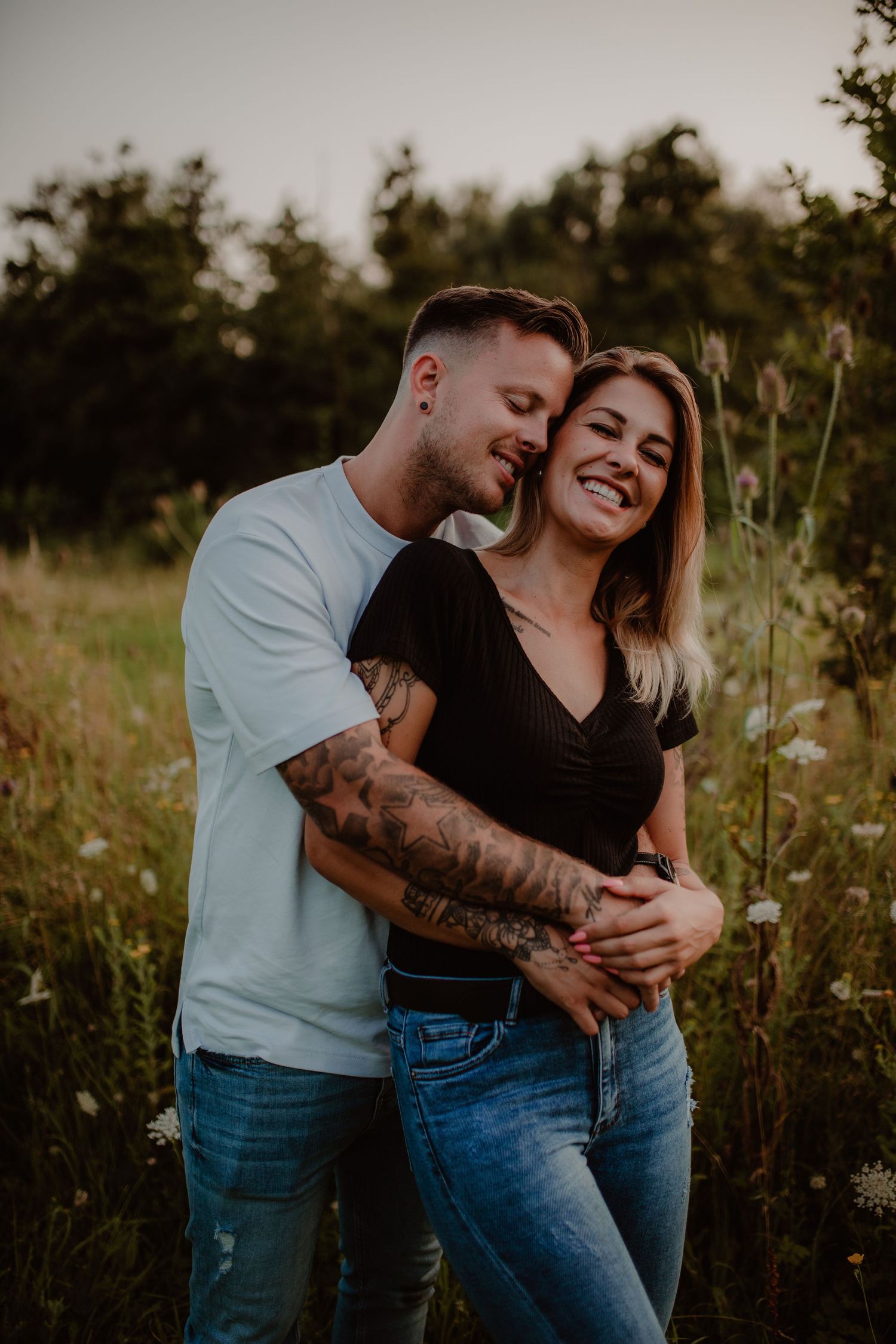 A romantic couple embraces in a wildflower field at sunset wearing jeans and casual attire.