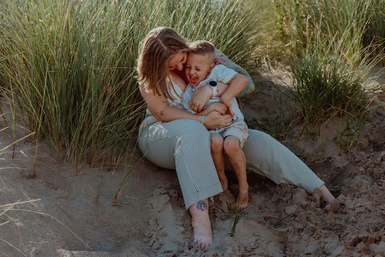 A mother and young child share a tender embrace while sitting on sandy beach dunes surrounded by tall grass.