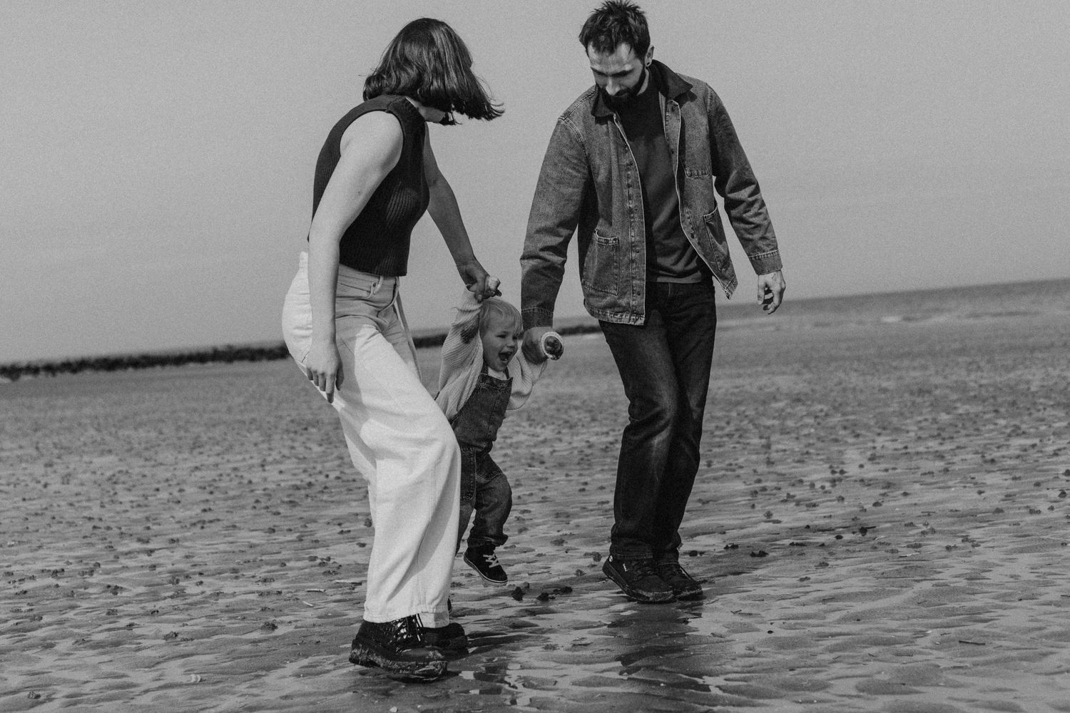 Family of three holding hands on a beach, black and white photo of parents swinging child over water.