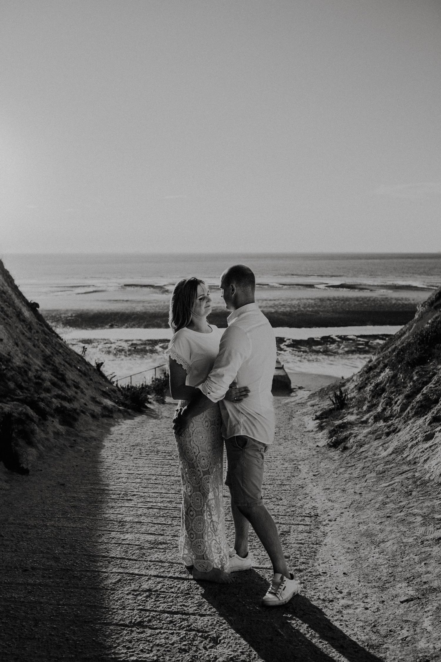 A couple embraces on a sandy path overlooking the ocean at sunset in black and white photography.