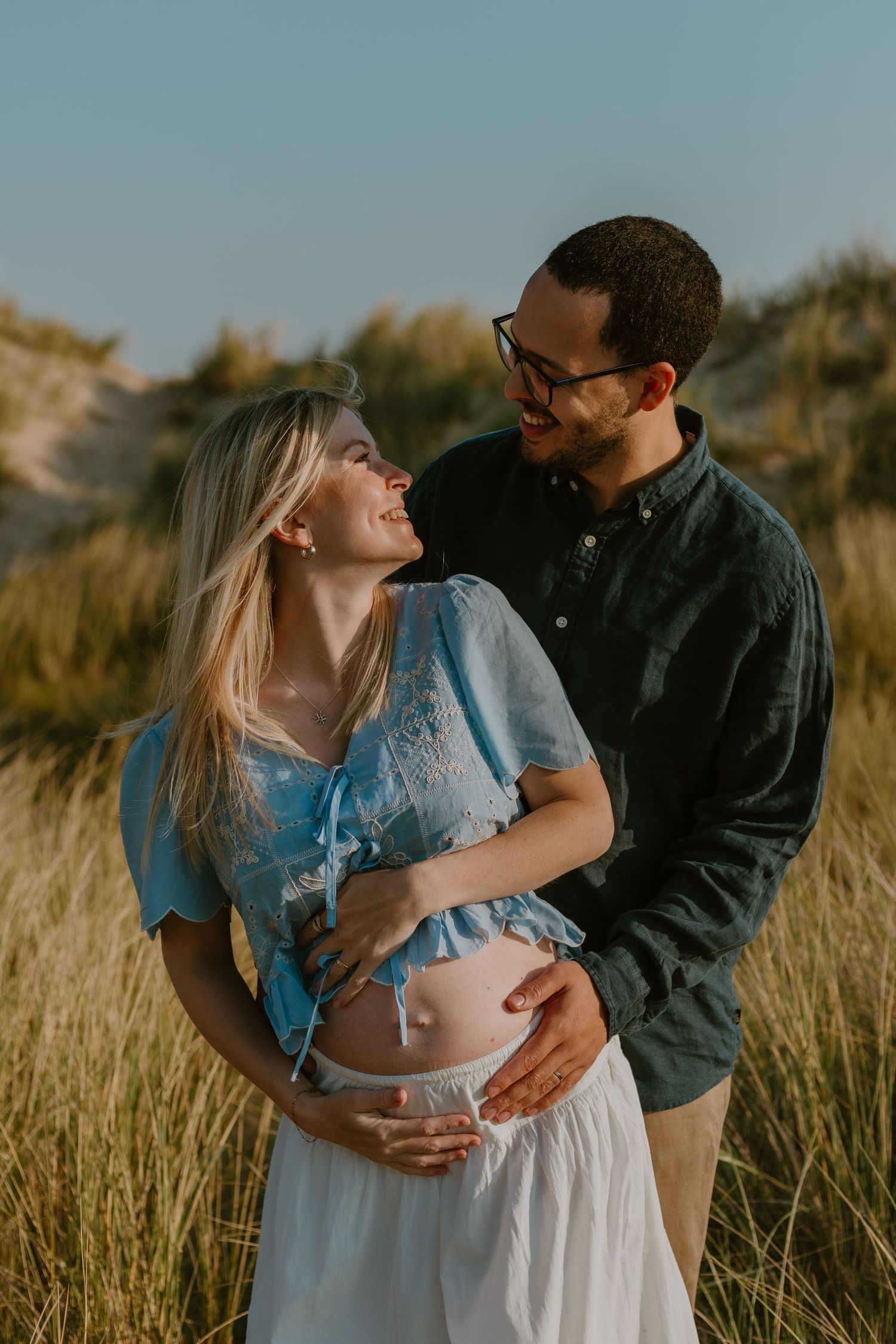 Smiling pregnant couple embracing outdoors in golden grass field during sunset maternity photo session.