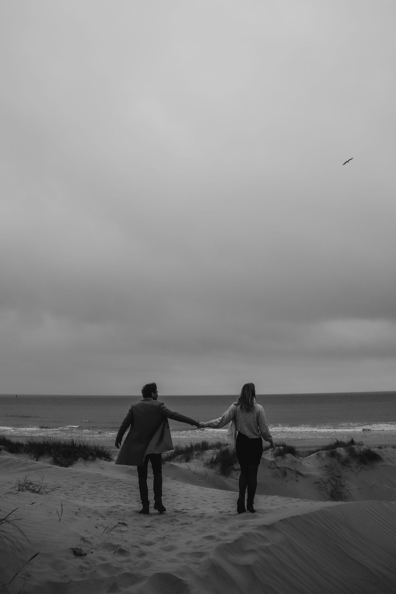 Silhouetten van twee mensen die hand in hand staan op het strand tegen een sombere grijze lucht.